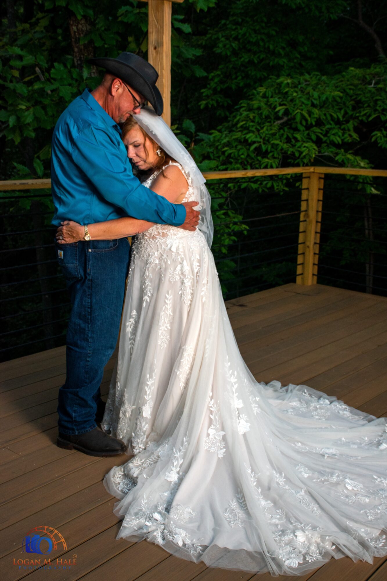 A man and woman embrace on a wooden deck with green trees in the background, the woman in a wedding dress.