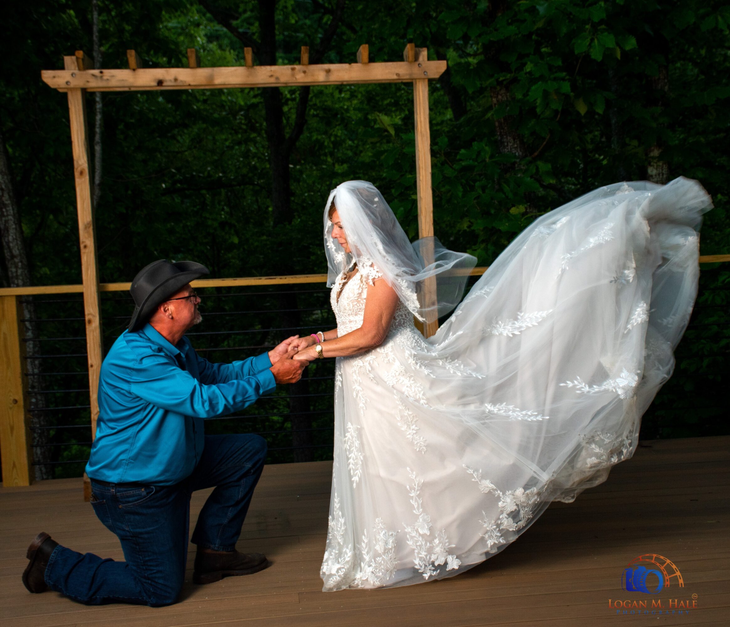 Person kneeling holding hands of a bride in a white wedding dress and veil outdoors, wooden frame in background.