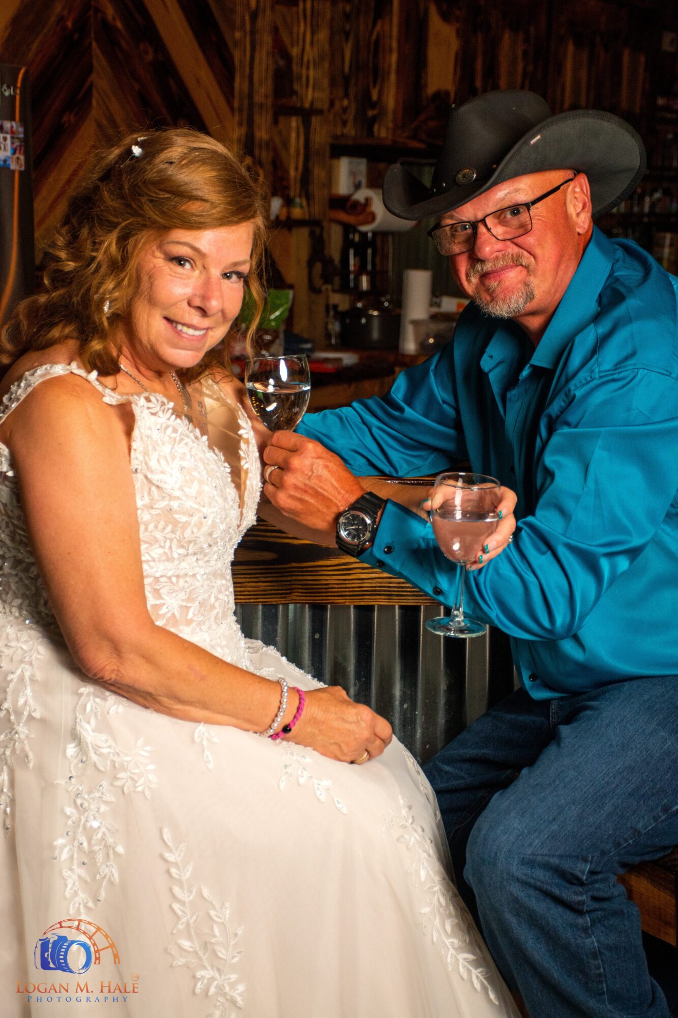 A woman in a white dress and a man in a cowboy hat and blue shirt sit together, smiling, with wine glasses in a rustic setting.