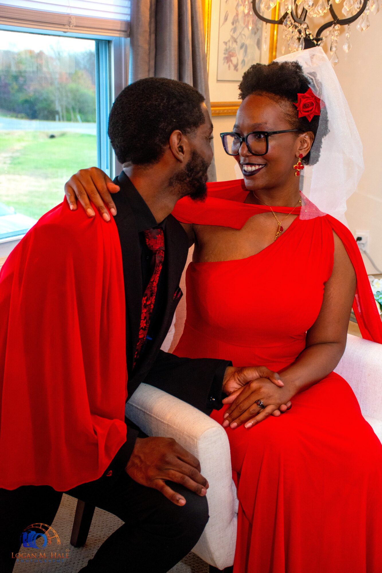 A couple dressed in red, sitting close and smiling indoors with a window and chandelier in background.