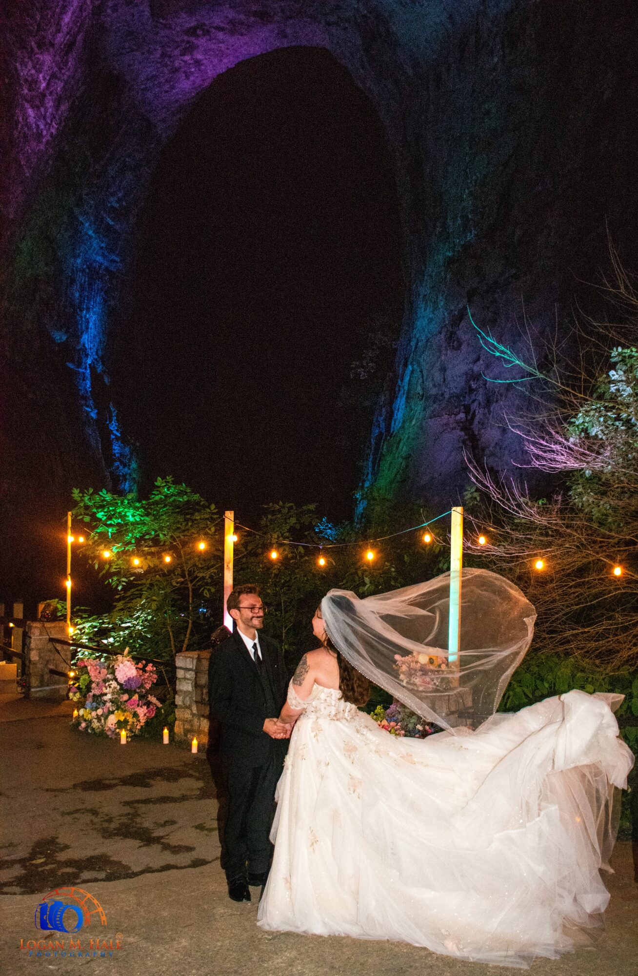 A bride and groom dance outdoors at night, with a large dark tunnel and string lights in the background.