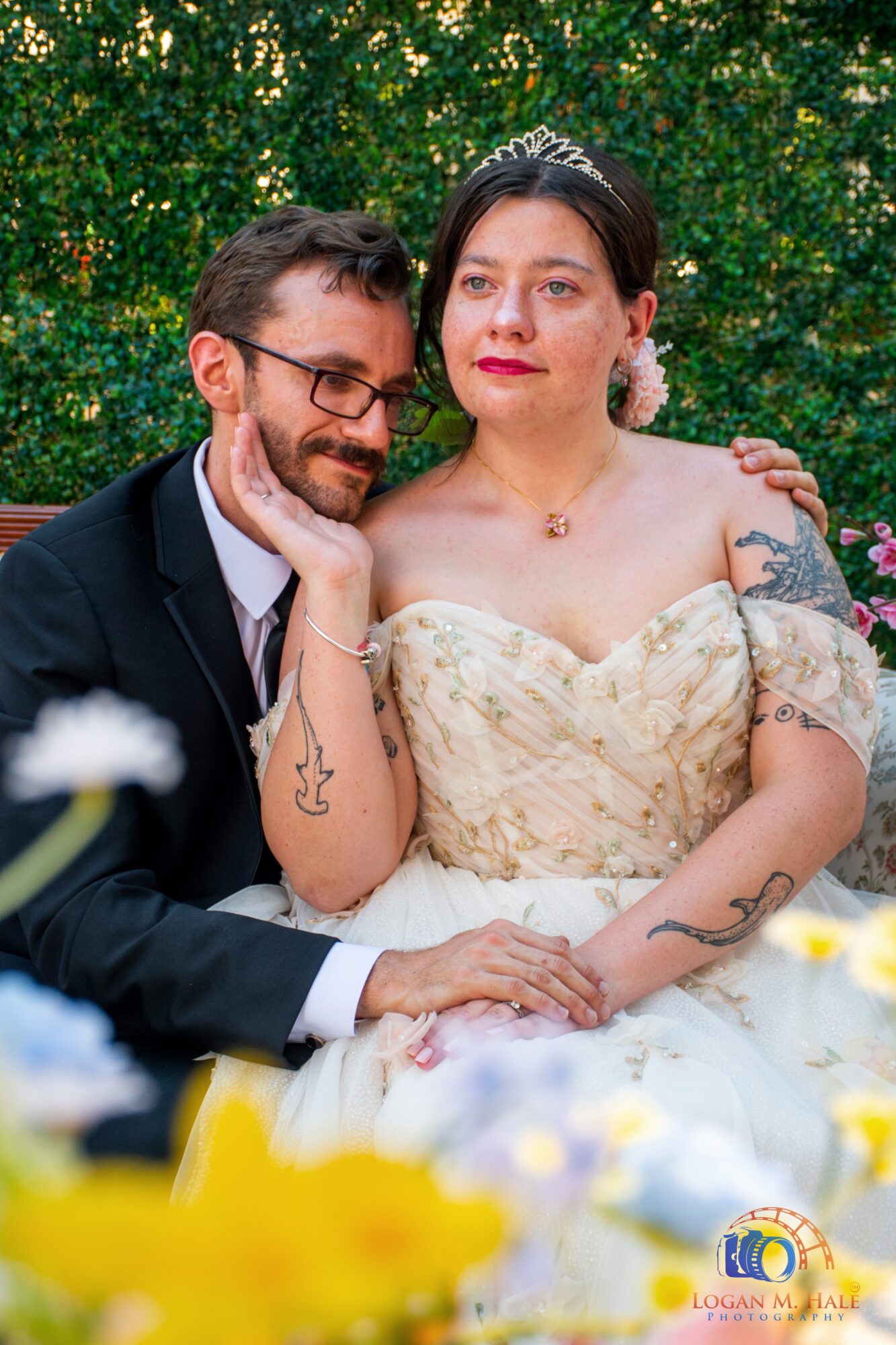A couple dressed in wedding attire sitting outdoors with greenery in the background, smiling and embracing.