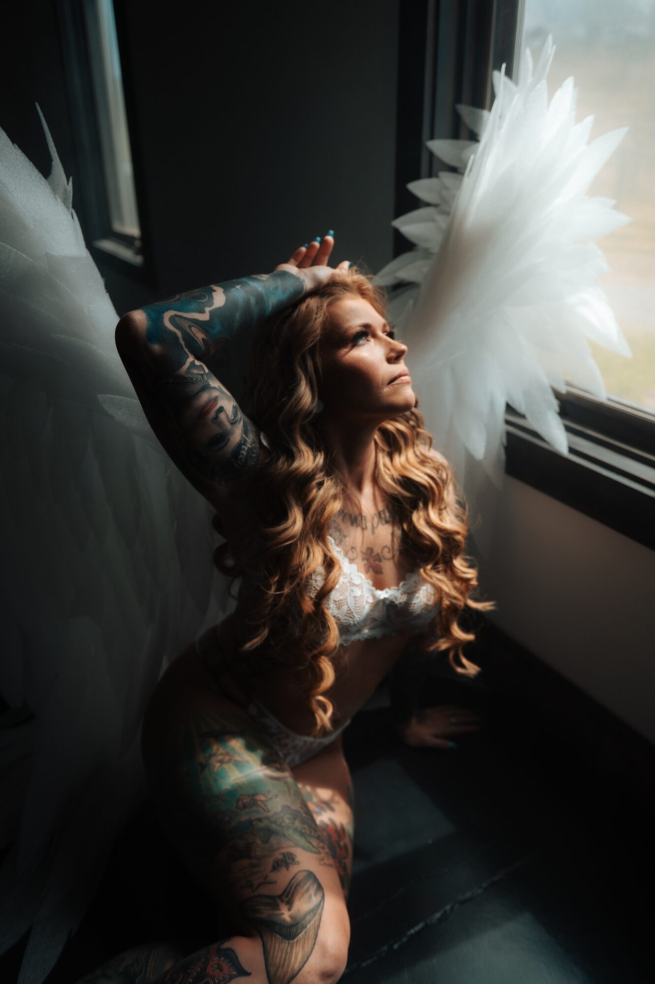 Woman with long hair and tattoos sitting by a window with angel wings, looking up, sunlight illuminating her face.