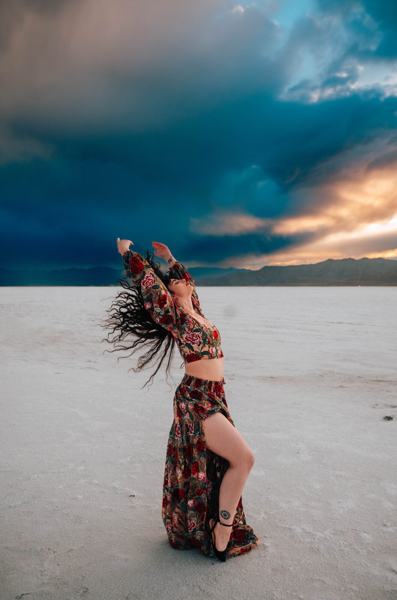 Person with long hair dancing on a sandy beach under a dramatic cloudy sky, wearing a floral outfit and high heels.
