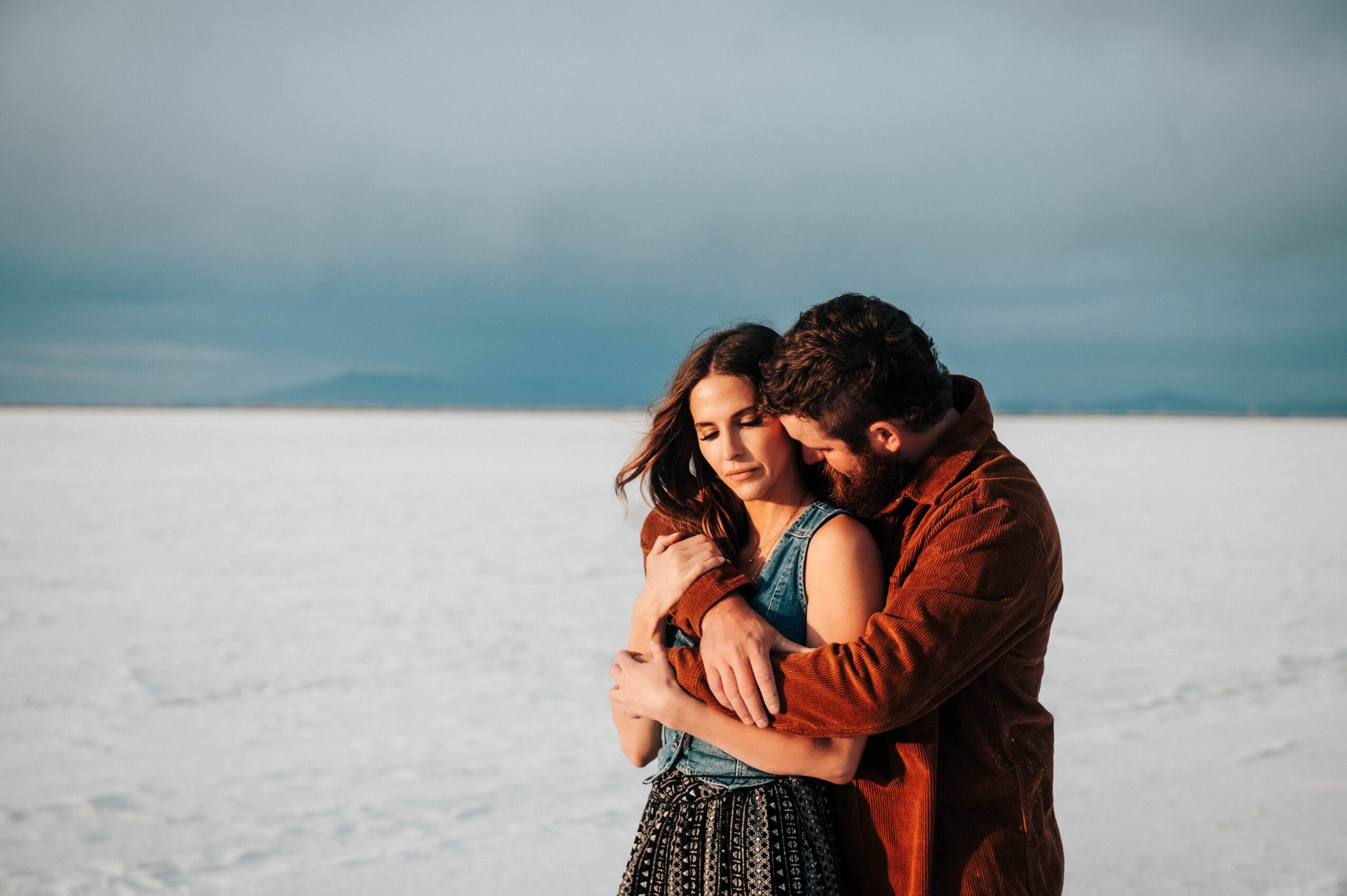 A couple embraces on a vast, flat, white landscape with a cloudy sky in the background.