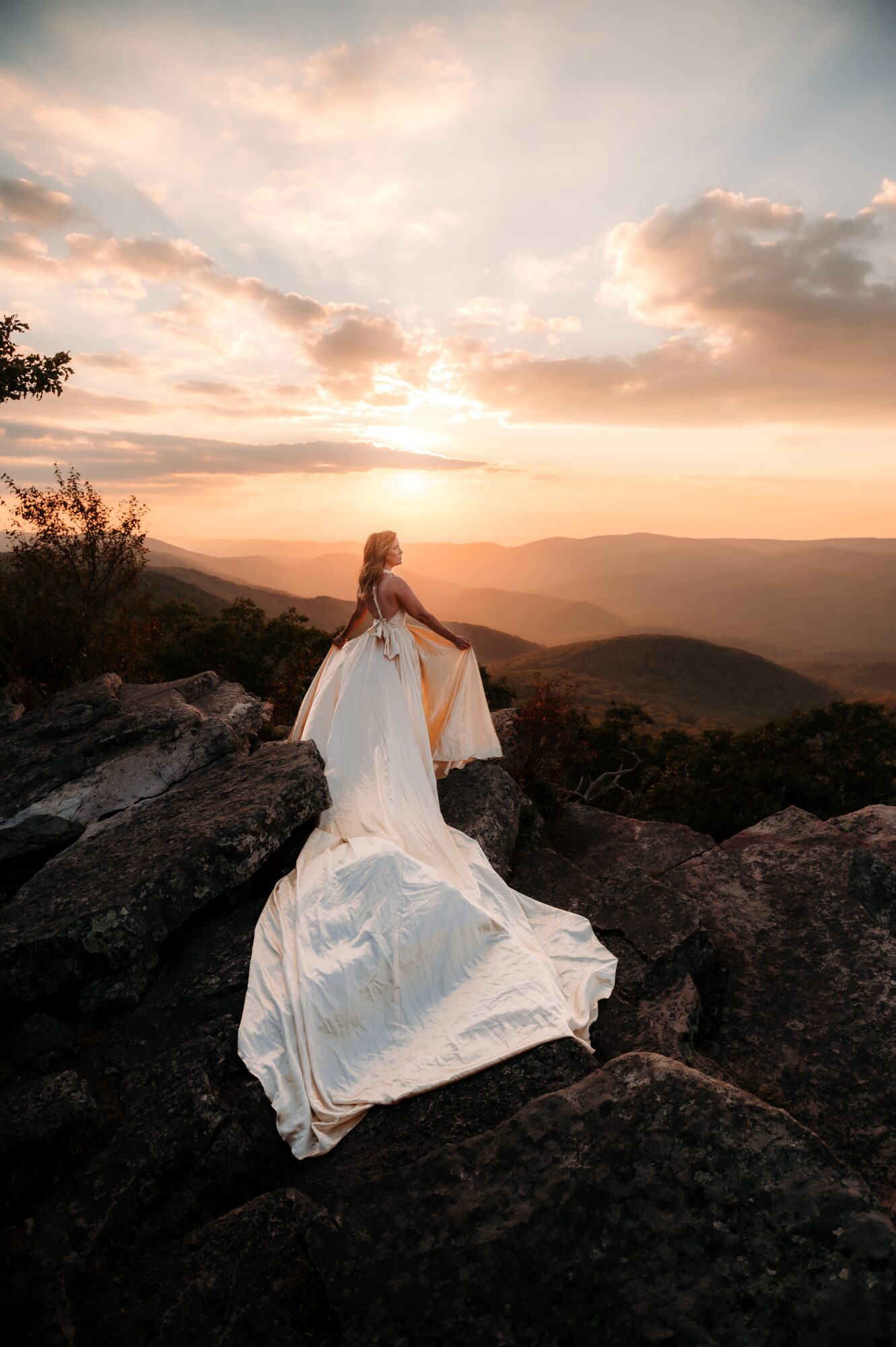 Woman in a white dress sitting on rocks overlooking a sunset landscape with clouds and distant mountains.