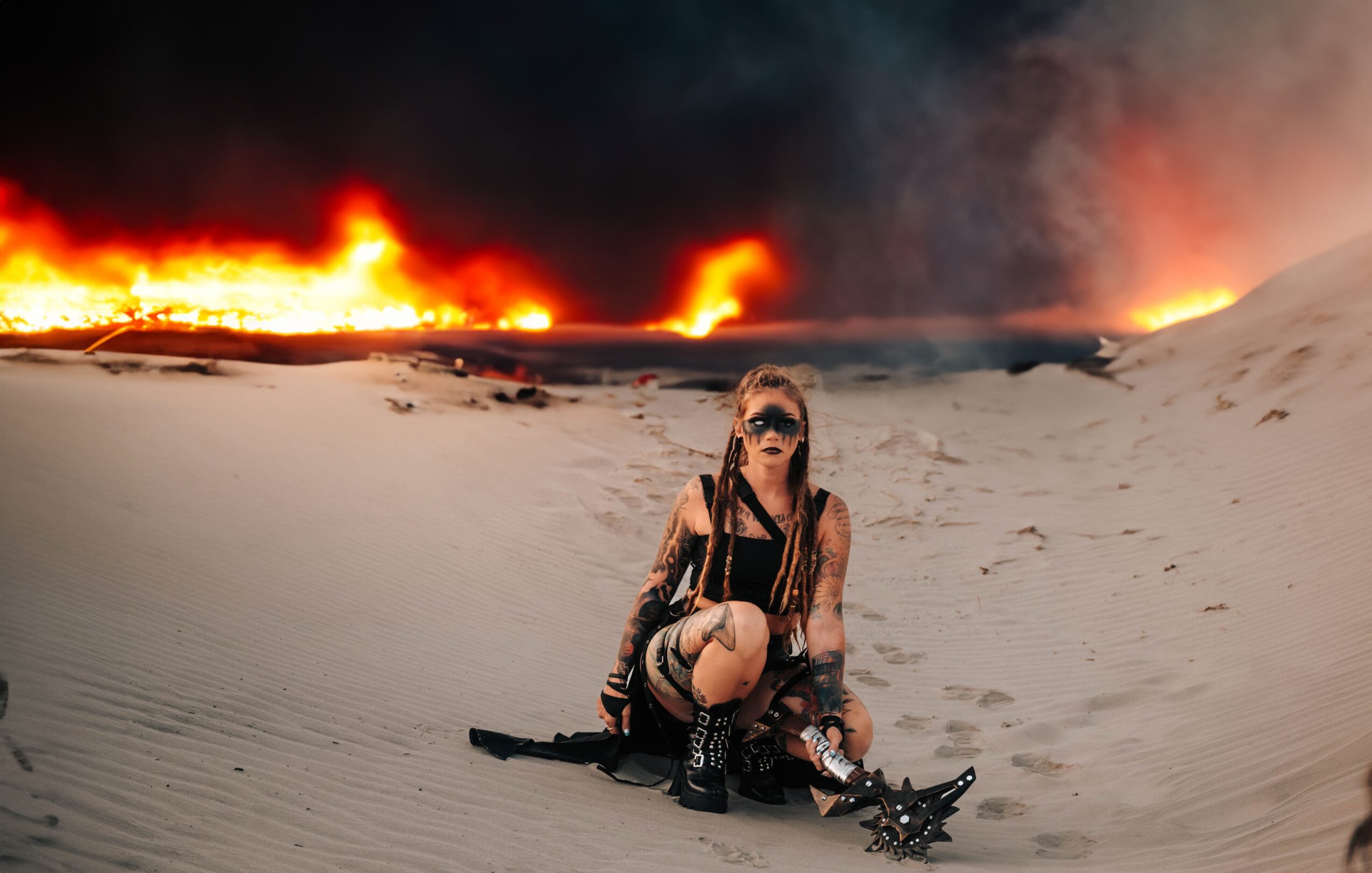Person sitting on sand with a fire and smoke in the background, wearing sunglasses and dark clothing.