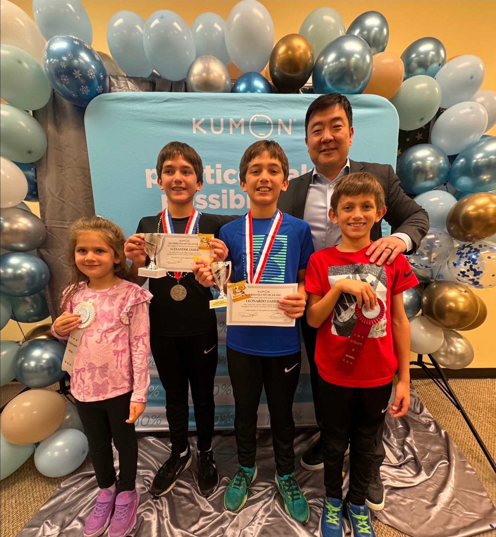 Group of five people, three children holding certificates and medals, with an adult, in front of balloon decorations and a blue backdrop.