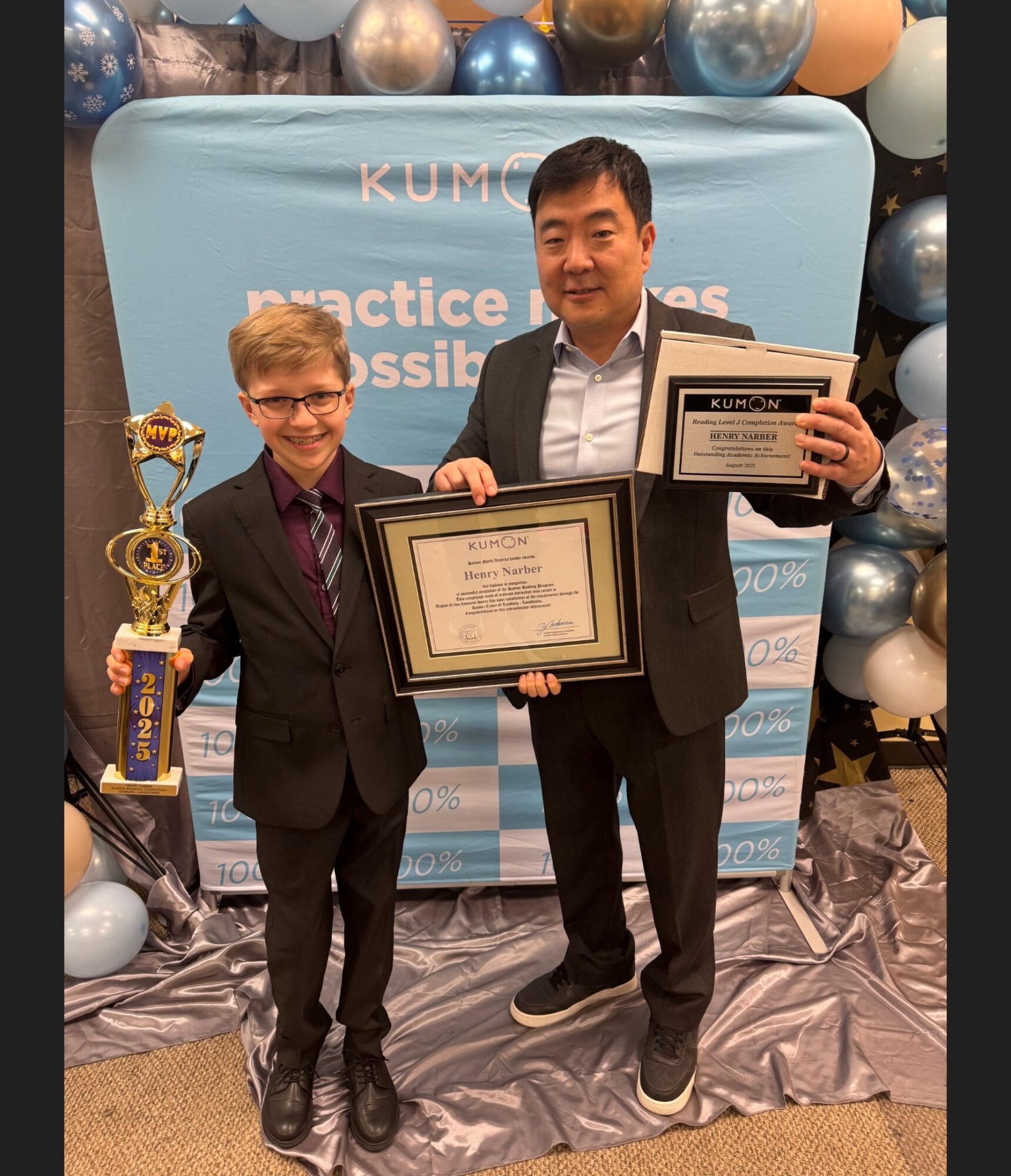 Two people in suits holding awards and certificates, standing in front of a decorated backdrop with balloons.