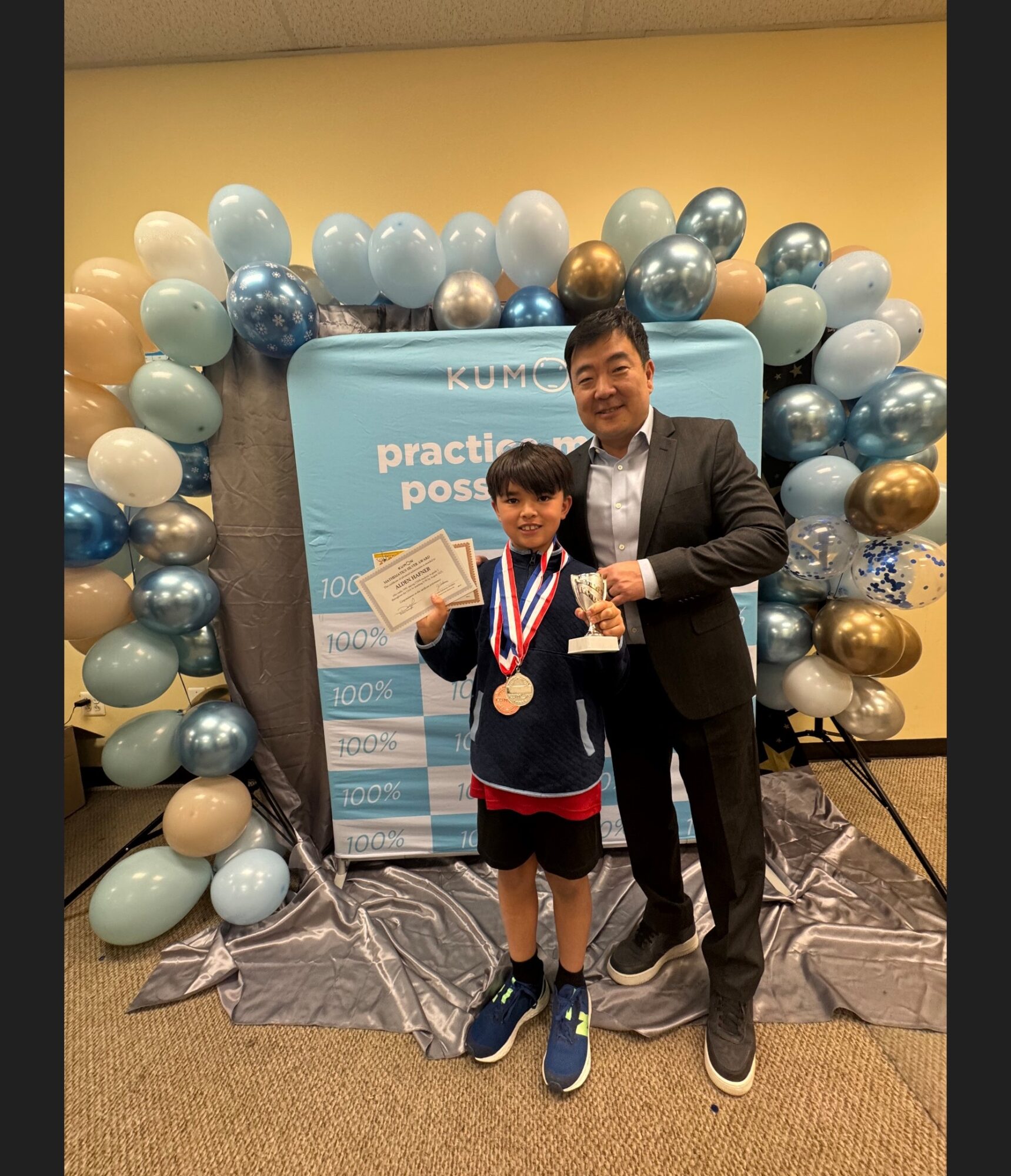 A young boy with medals and certificates stands next to a man in a suit, both smiling, in front of a balloon arch and backdrop.