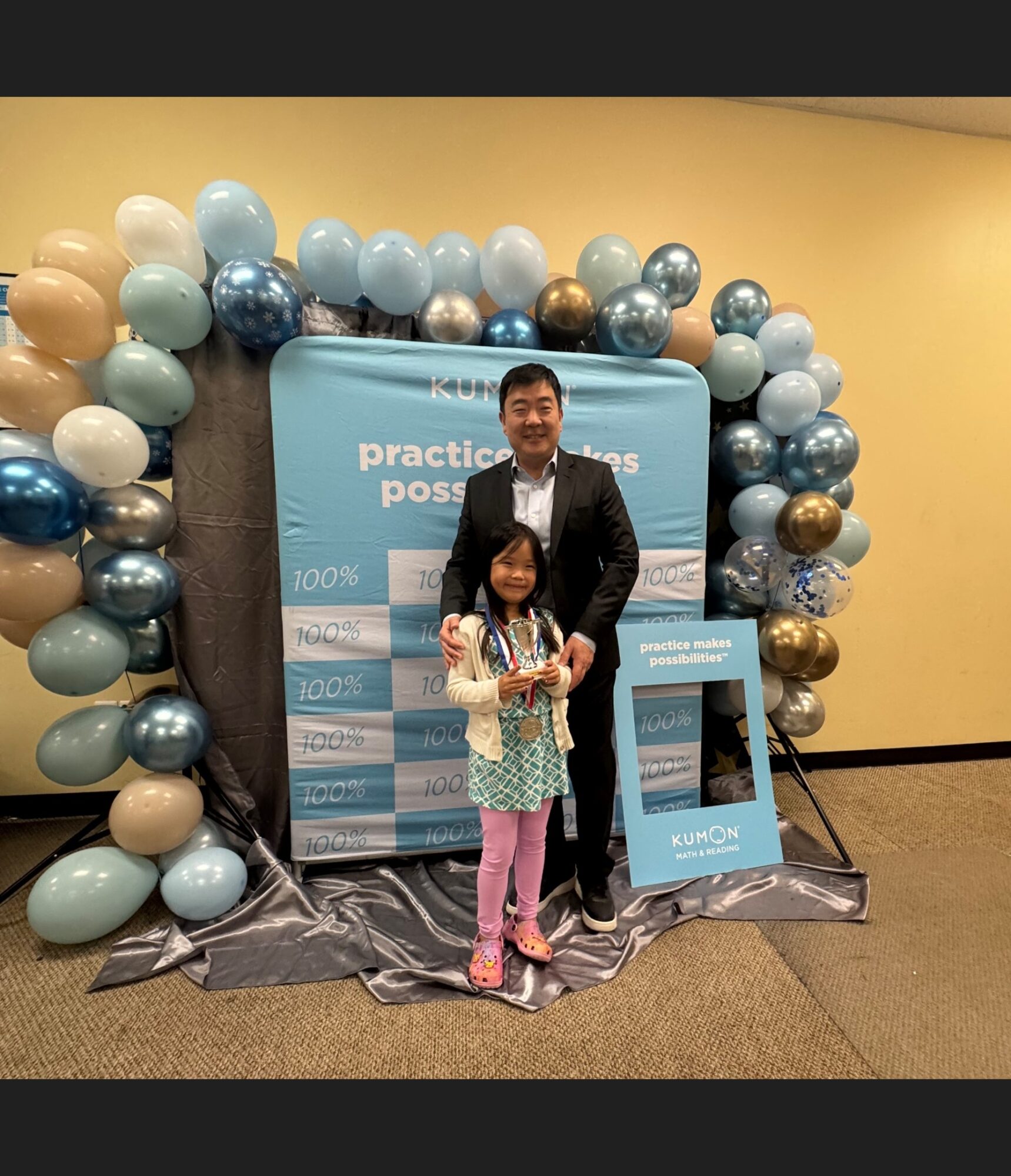 A man and a girl stand in front of a balloon arch and a large blue sign at an indoor event, smiling.