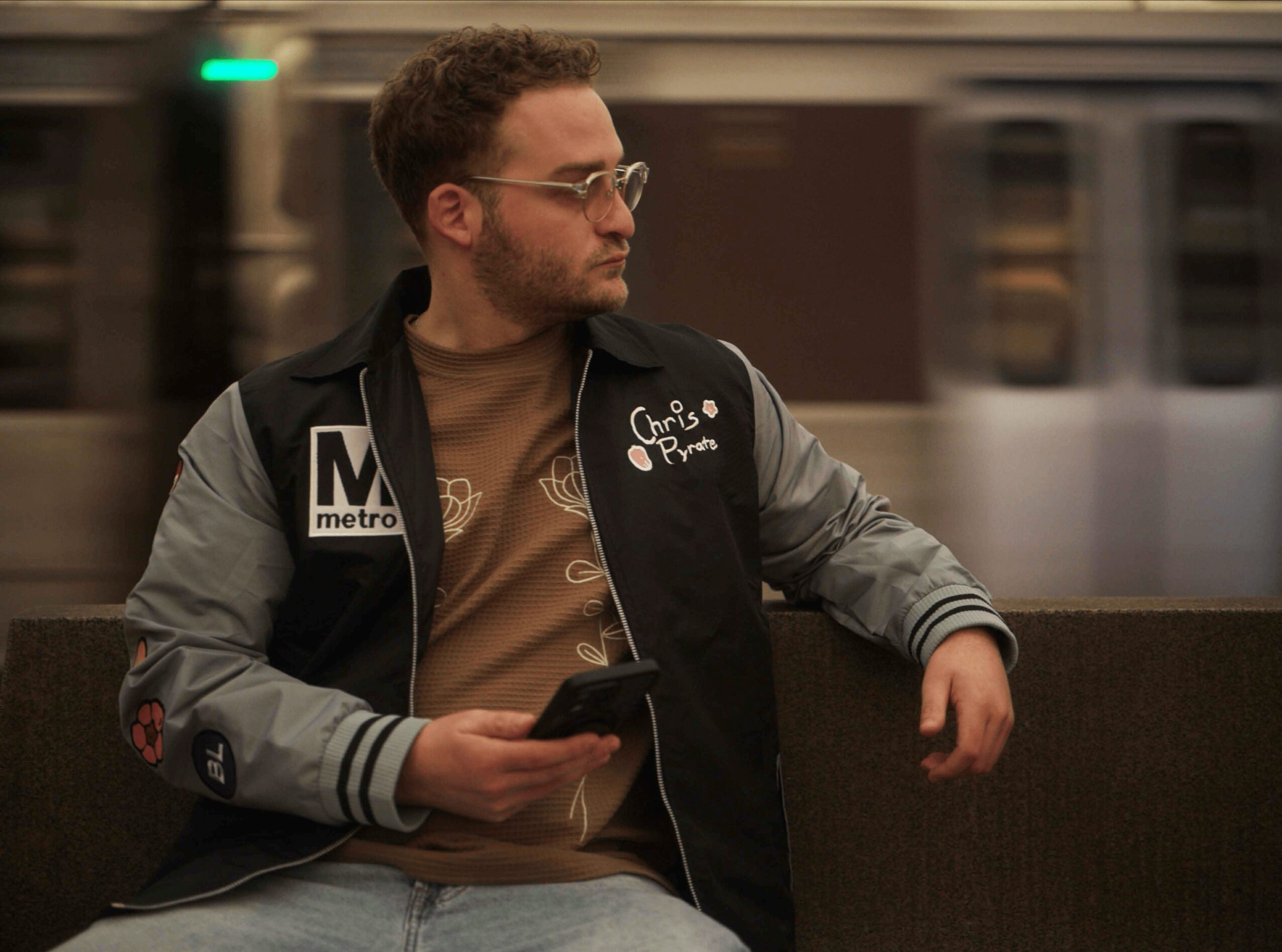 Man with glasses and beard sitting on a bench in a subway station, holding a phone, wearing a black and gray jacket.