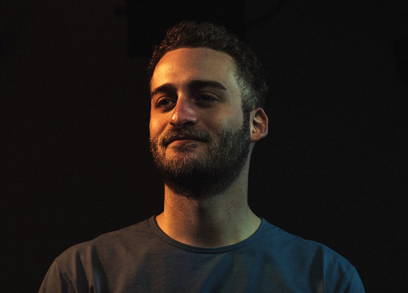 Man with dark curly hair and beard, wearing a dark shirt, looking to the side, dim background