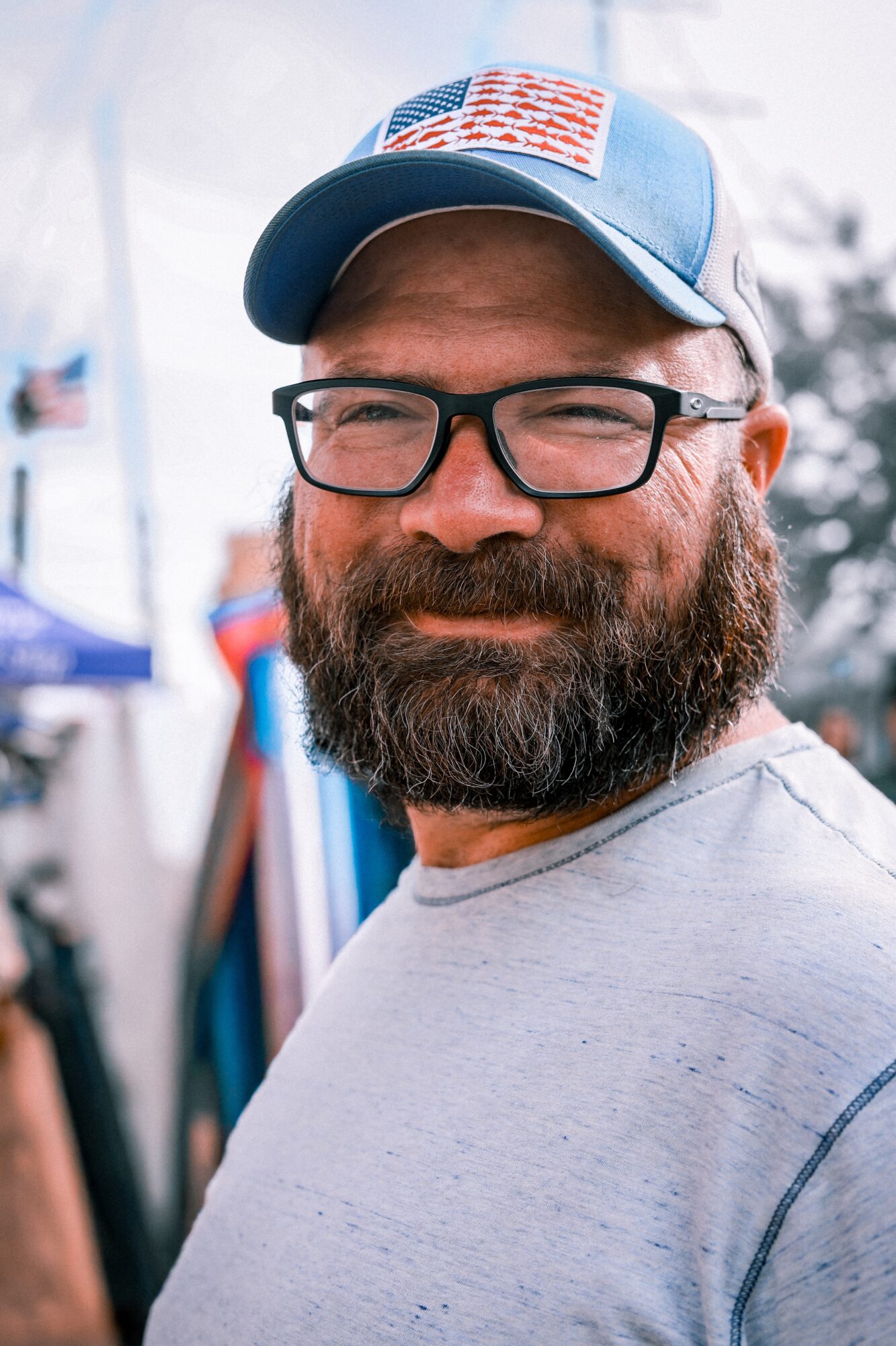 Man with glasses and a beard wearing a blue cap with an American flag patch, smiling outdoors.