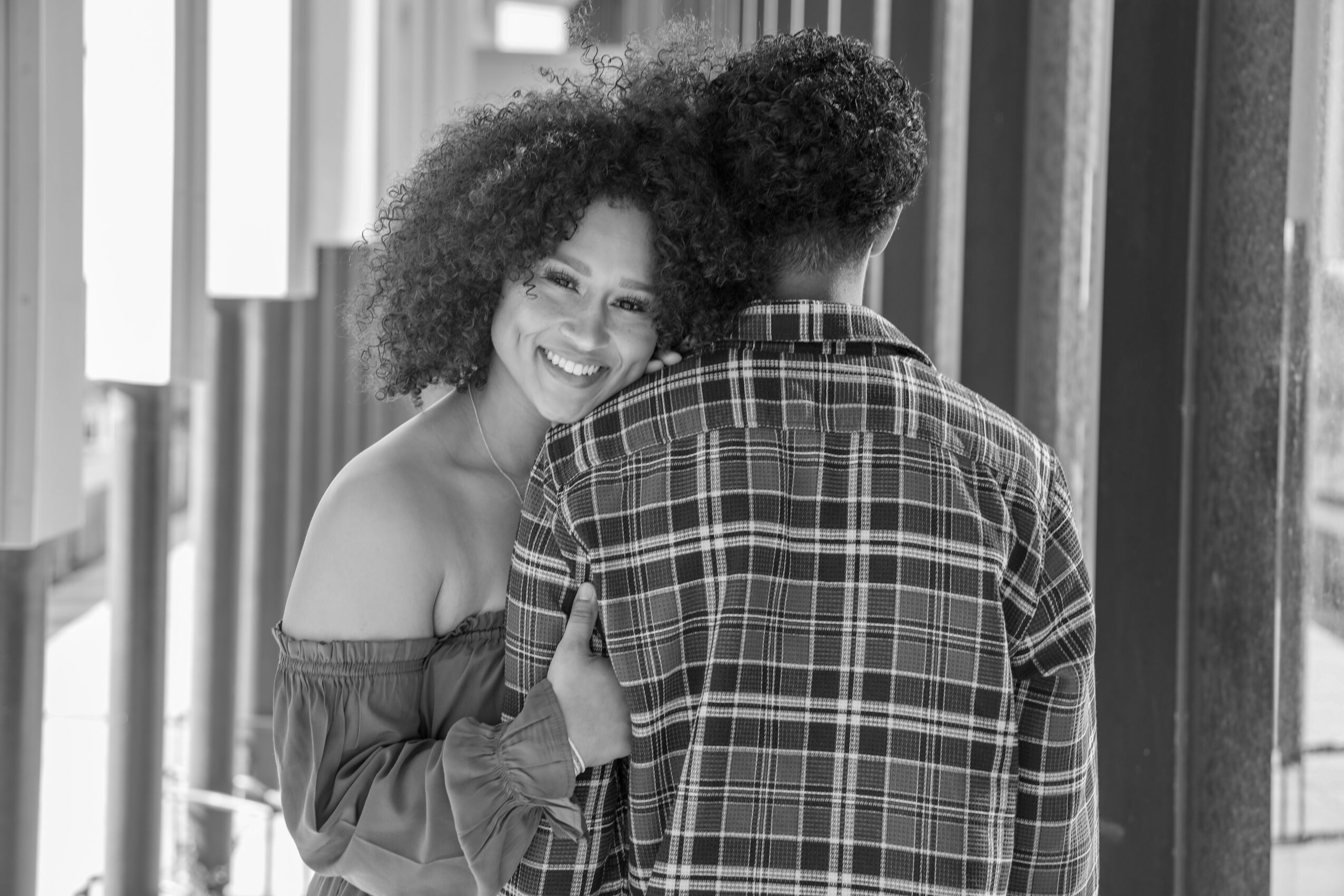 Smiling woman with curly hair leaning on a person with short hair in a plaid shirt, standing near windows.