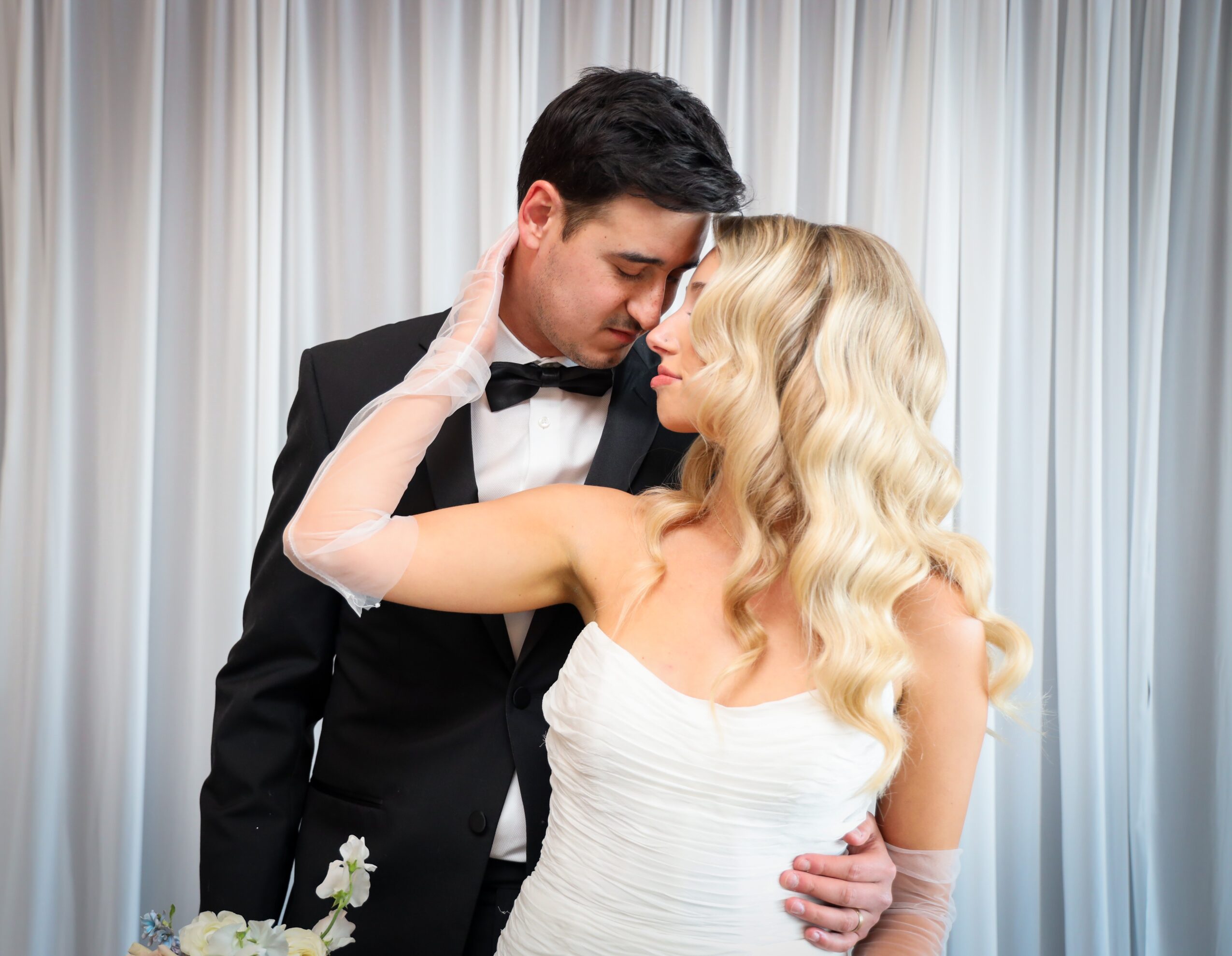 A couple in formal attire, the man in a tuxedo and the woman in a white dress, close together with their eyes closed.