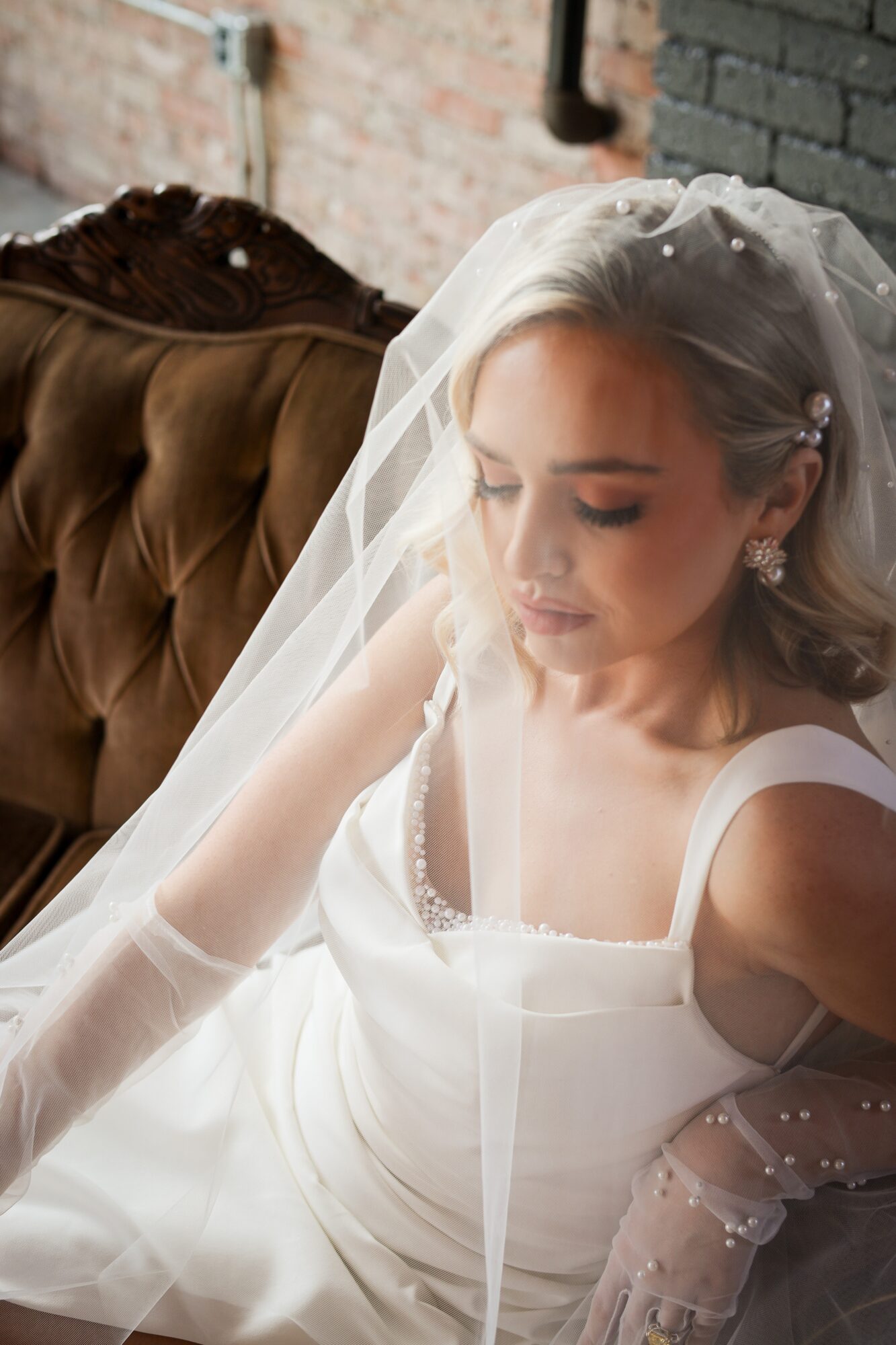 Bride with veil and white dress sitting on a vintage sofa, looking down, in a rustic setting.