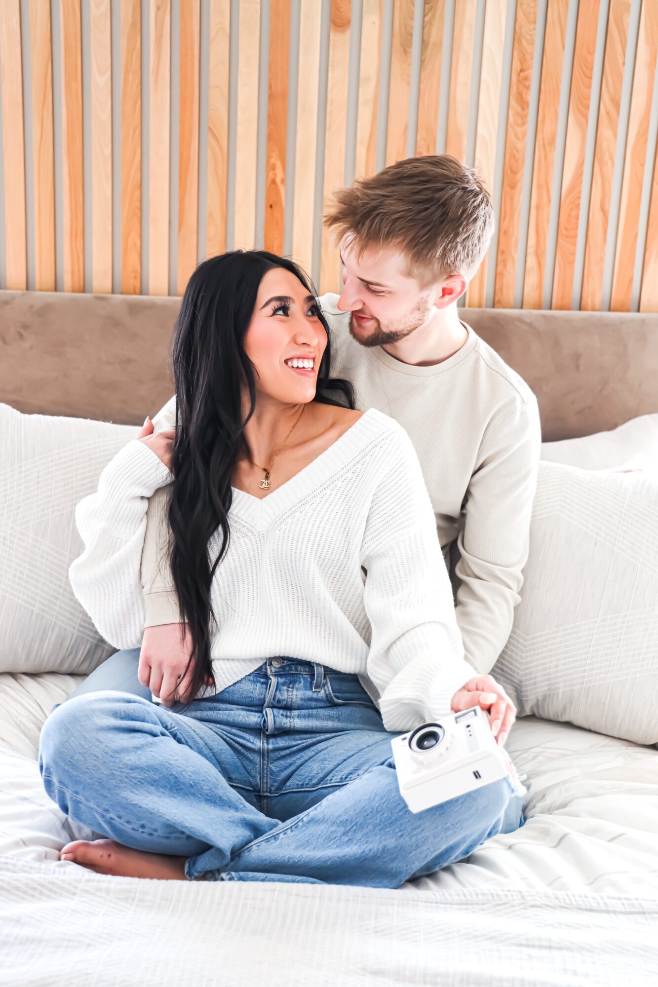A smiling woman with long dark hair and a man with short light hair sit on a bed, looking at each other.
