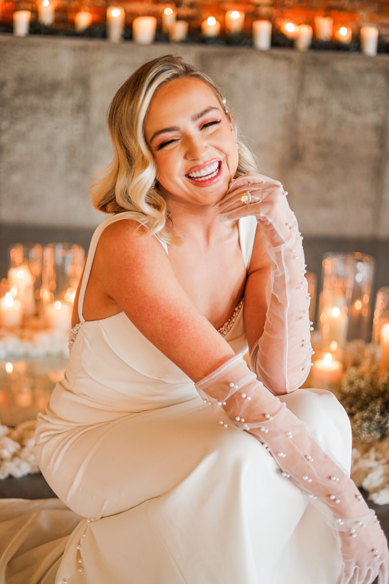 Smiling woman in a white dress with sheer gloves, sitting in front of candles, laughing with hand near face.