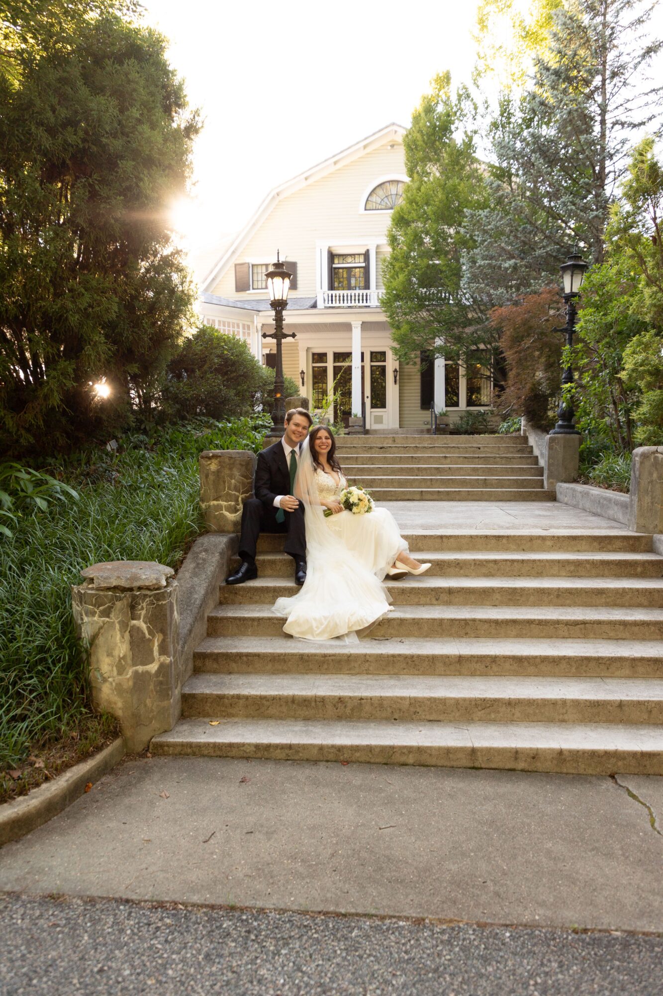 A bride and groom sit on steps outside a large white house surrounded by trees, sunlight shining through the foliage.