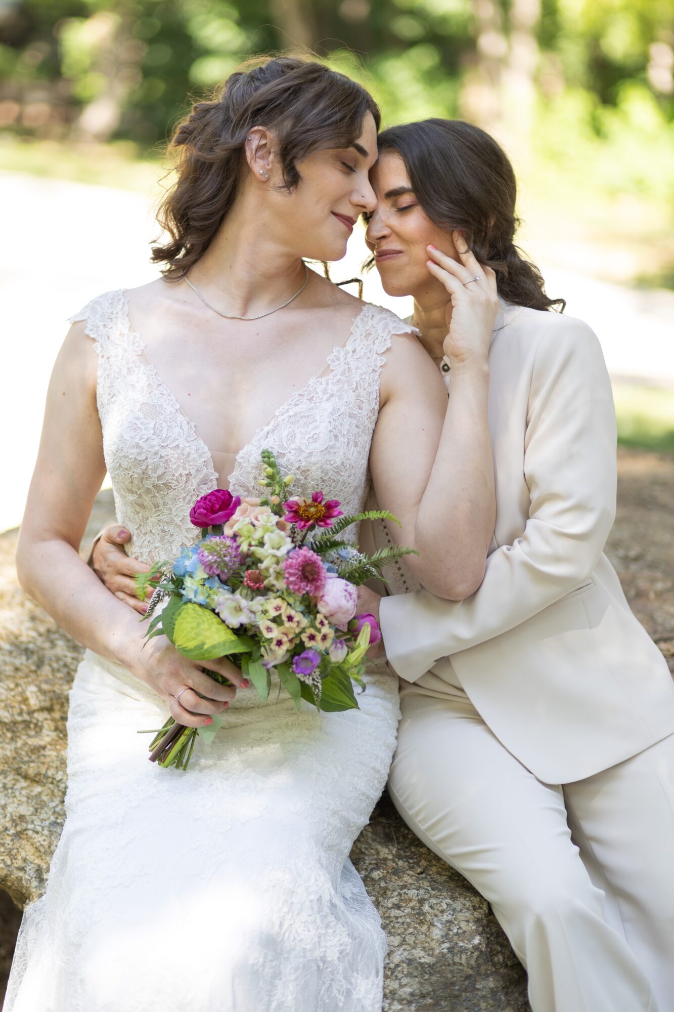 Two women in wedding attire sit close, one holding a bouquet of colorful flowers, outdoors with trees in background.