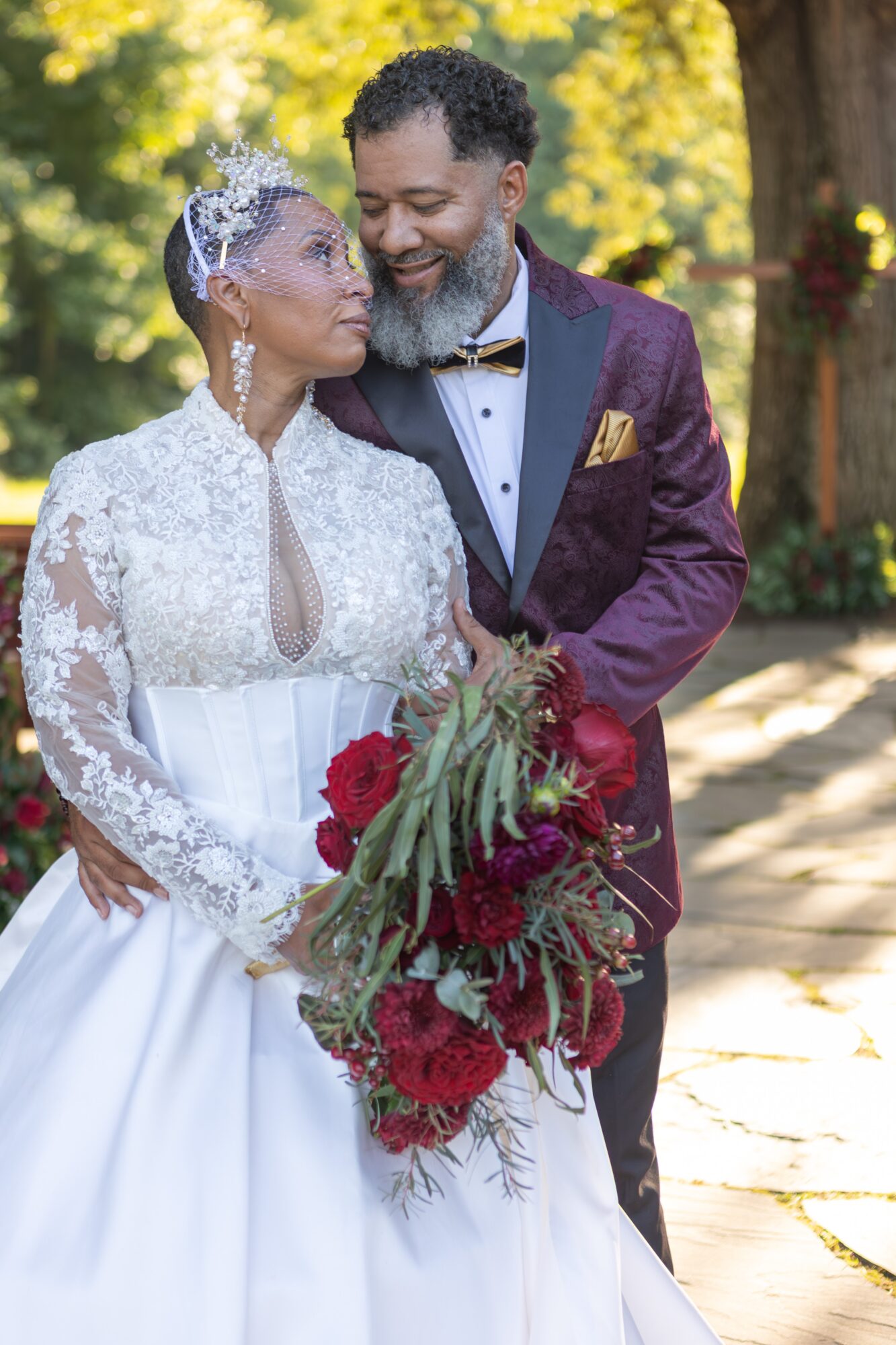 A bride and groom stand close outdoors, holding a bouquet of red flowers, smiling at each other.