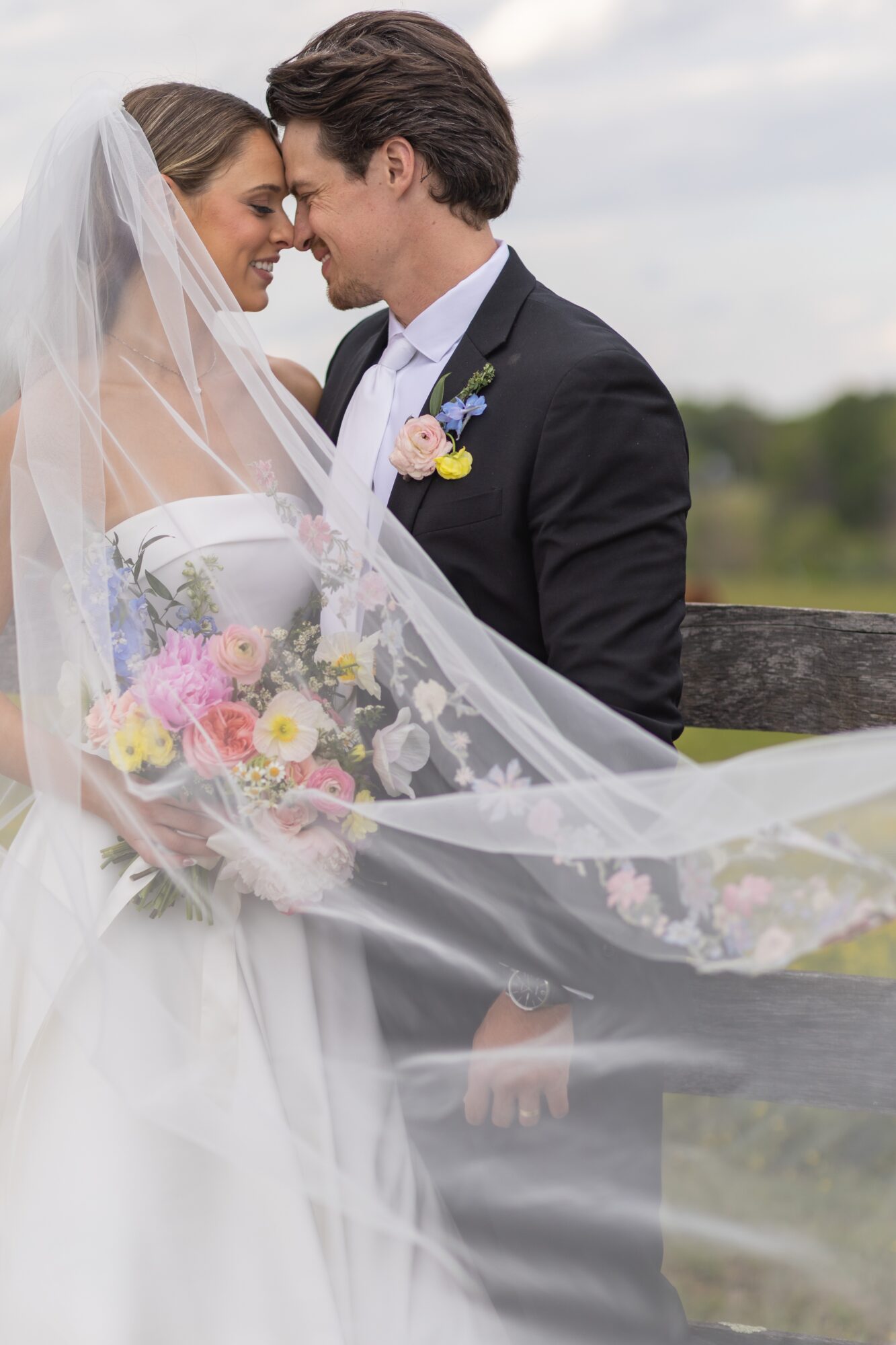 Bride and groom face each other outdoors, smiling, with veil and bouquet, near a wooden fence and water background.