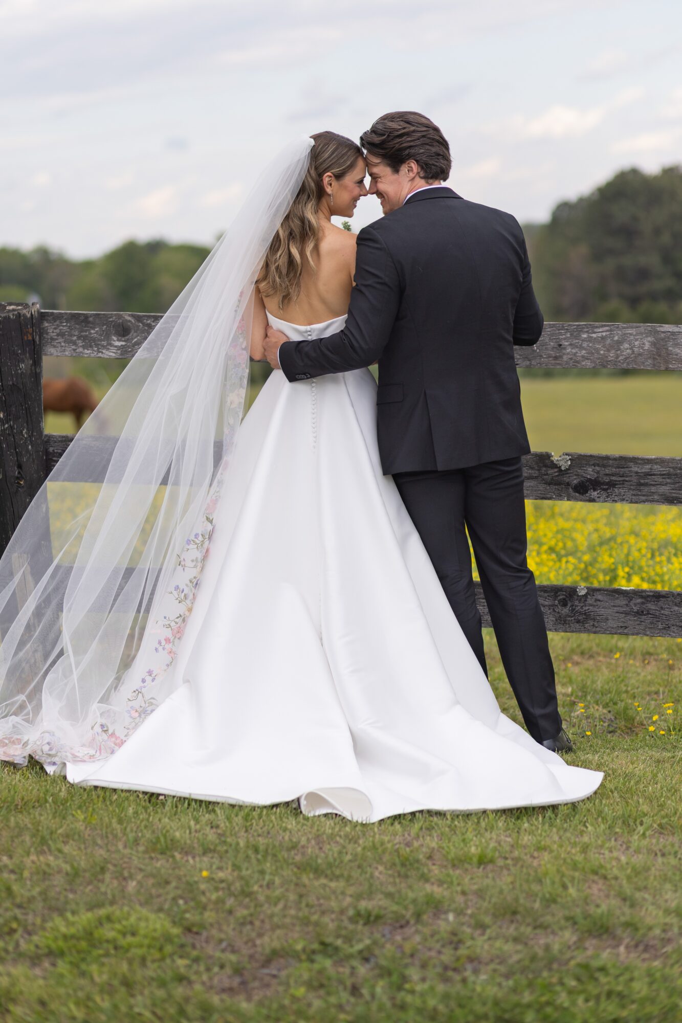 Bride and groom stand close together outdoors, facing each other, with a wooden fence and green landscape in the background.