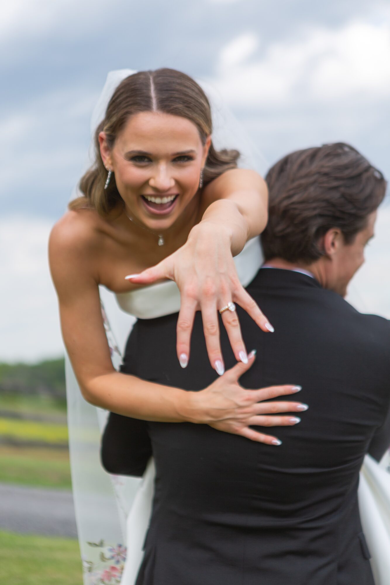 Woman in wedding dress showing her ring, smiling, with a man in a suit outdoors under a cloudy sky.