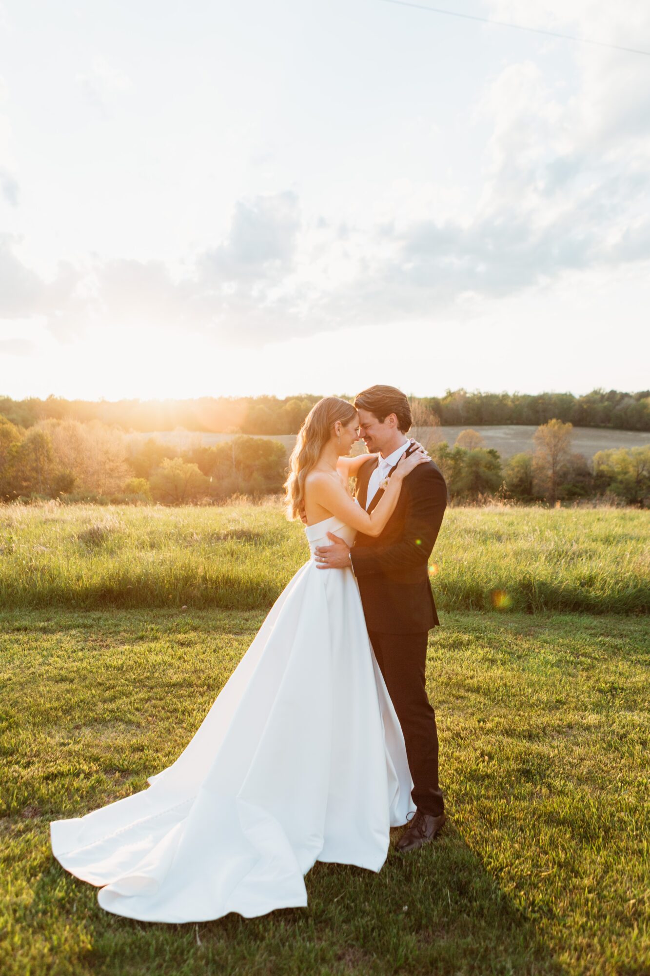 A bride and groom stand close together in a field during sunset, embracing and looking at each other.