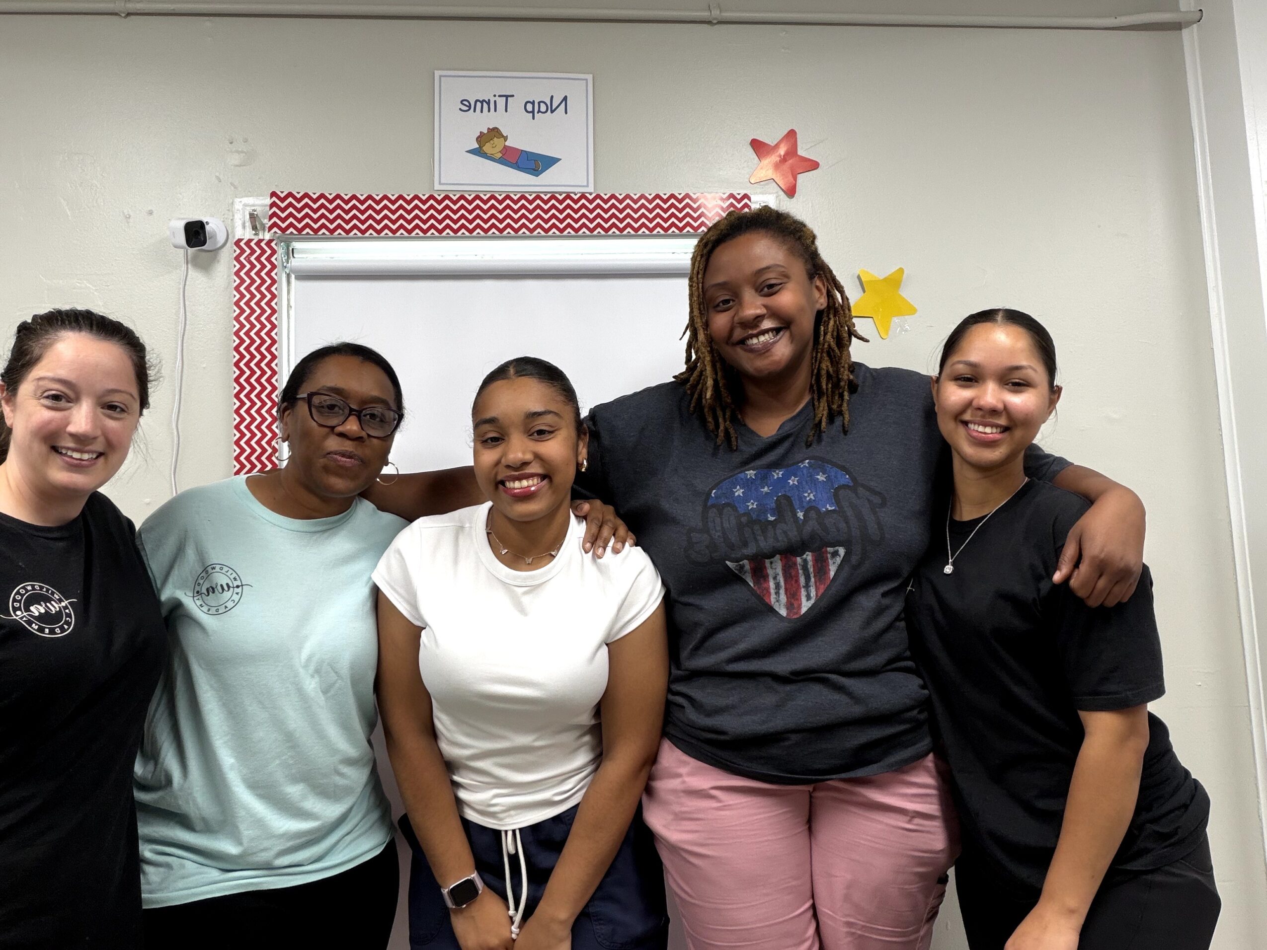 Five smiling women standing together in a classroom, with a whiteboard and colorful decorations behind them.