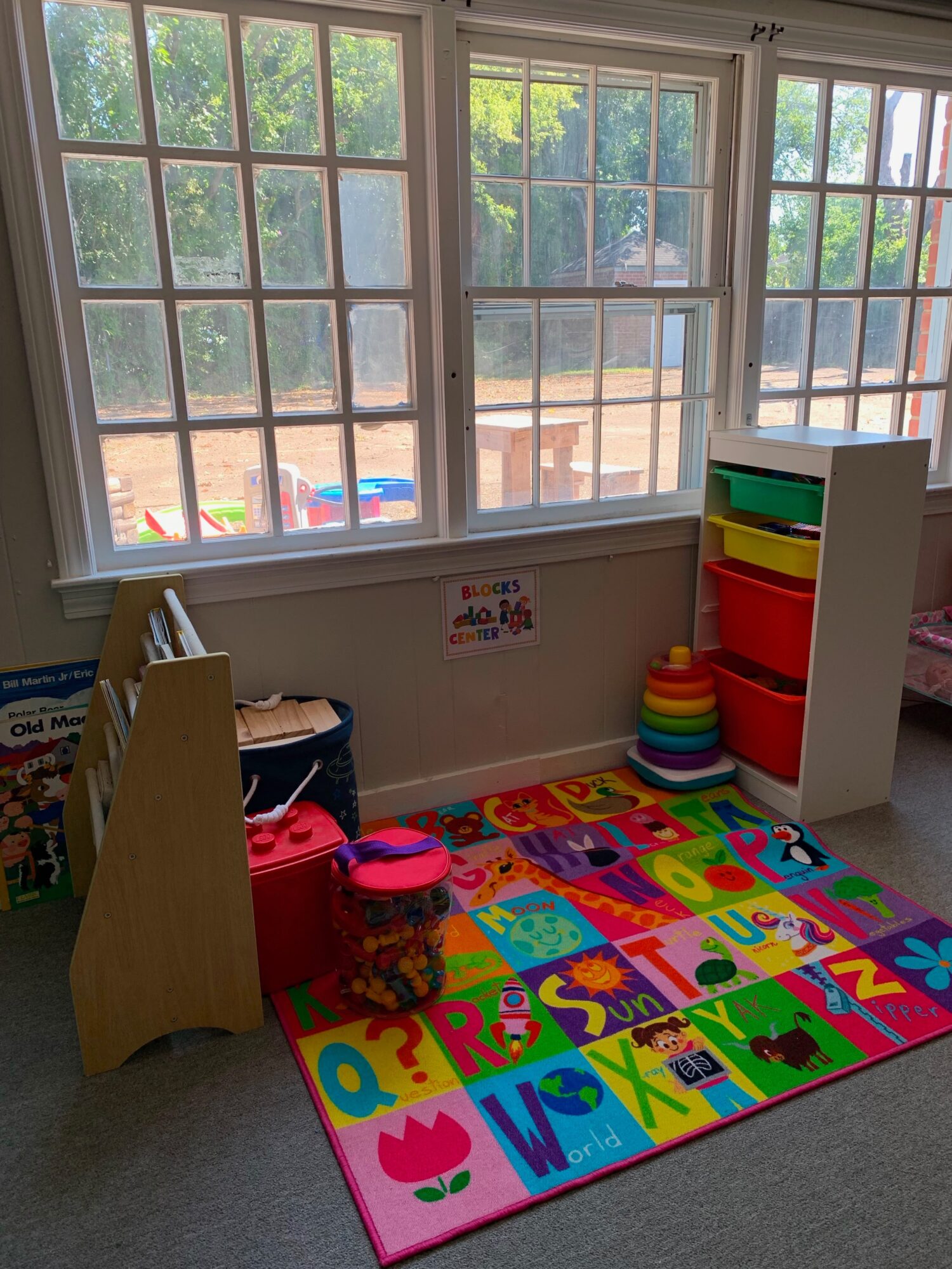 Children's play area with colorful foam alphabet mat, stacked rings, and a small bookshelf near large windows showing outdoor trees.