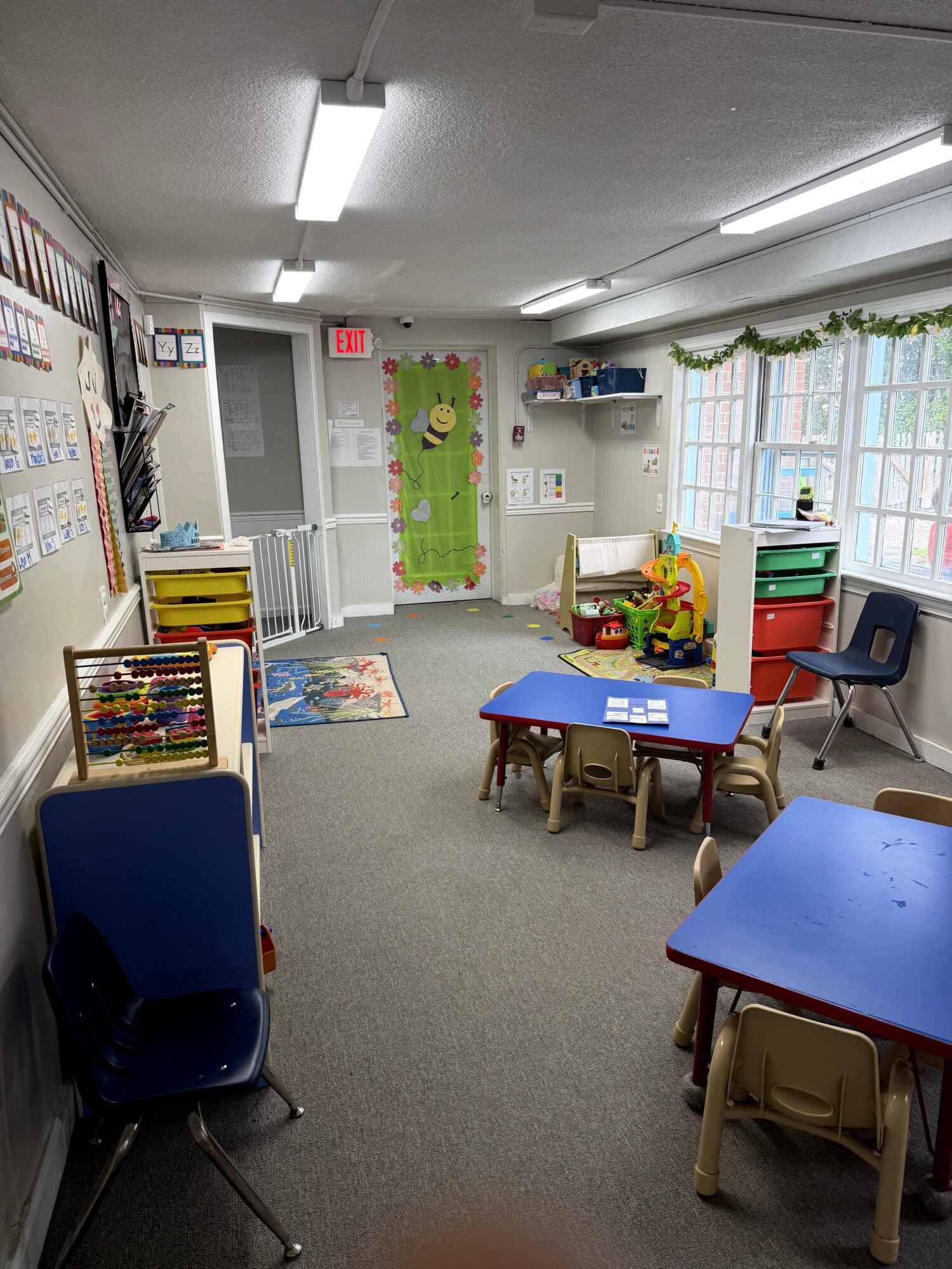 Classroom with tables, chairs, and educational decorations, including a green wall hanging and window with curtains.