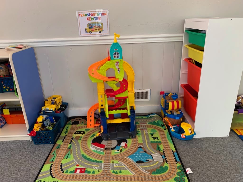 Colorful toy train on a play mat with roads and buildings, surrounded by toy vehicles and storage shelves in a playroom.