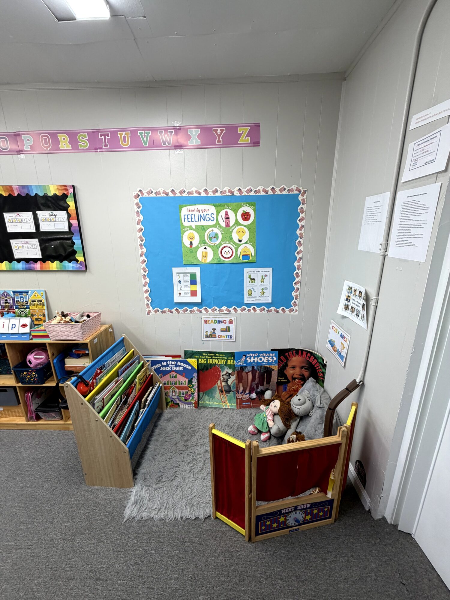 Corner of a classroom with a bookshelf, a puppet sitting in a small chair, and a colorful bulletin board.