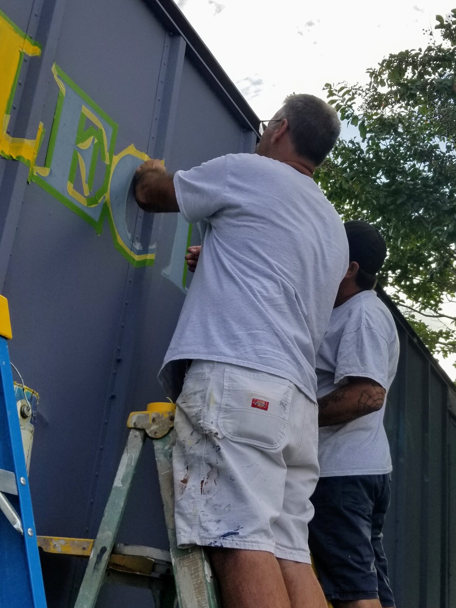 Two men painting a large blue wall with yellow and green letters outdoors, one standing on a ladder, trees in background.
