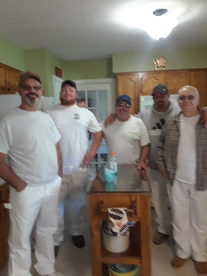 Five people standing together in a kitchen, smiling, with wooden cabinets and a table in the background.