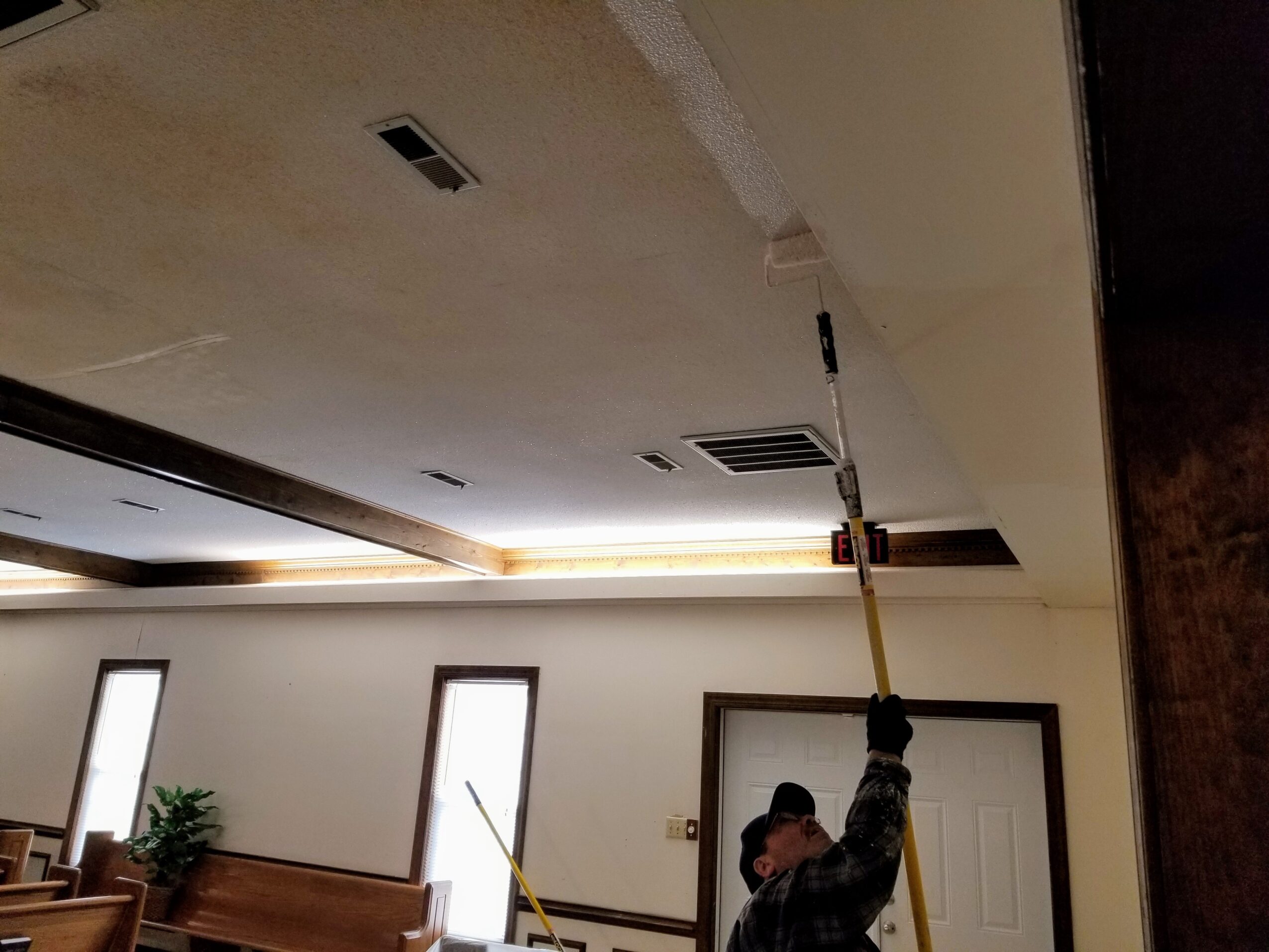 Person painting ceiling with a roller, room with windows and a door, ceiling partially painted, indoor setting.