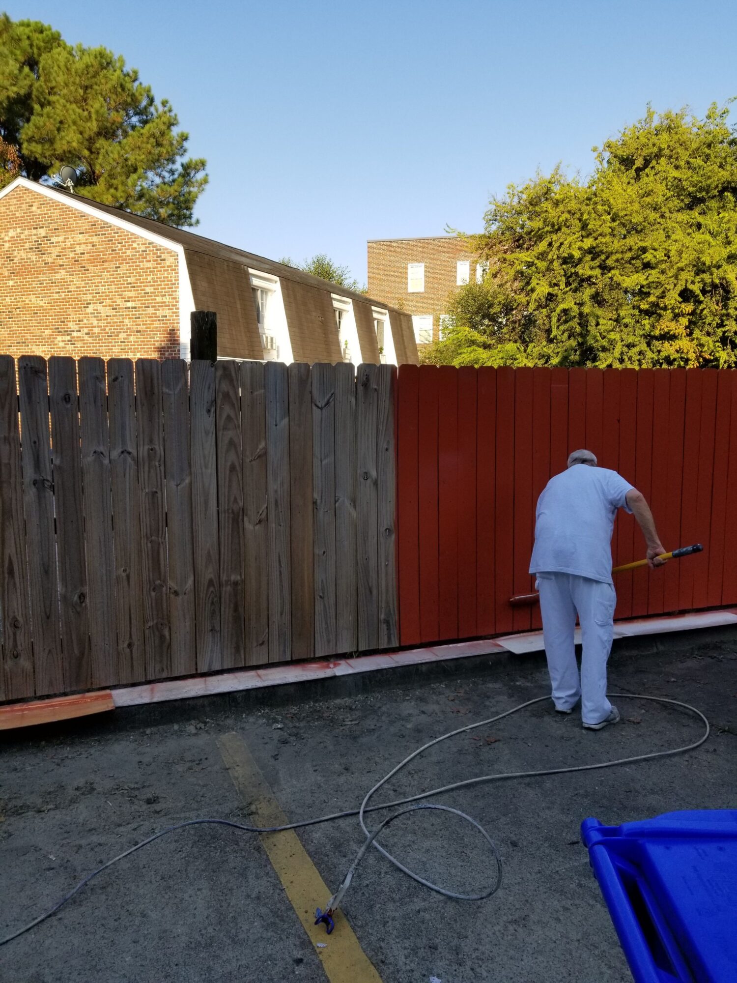 Person in white clothing cleaning a red fence with a hose, outdoor setting, trees and building in background.
