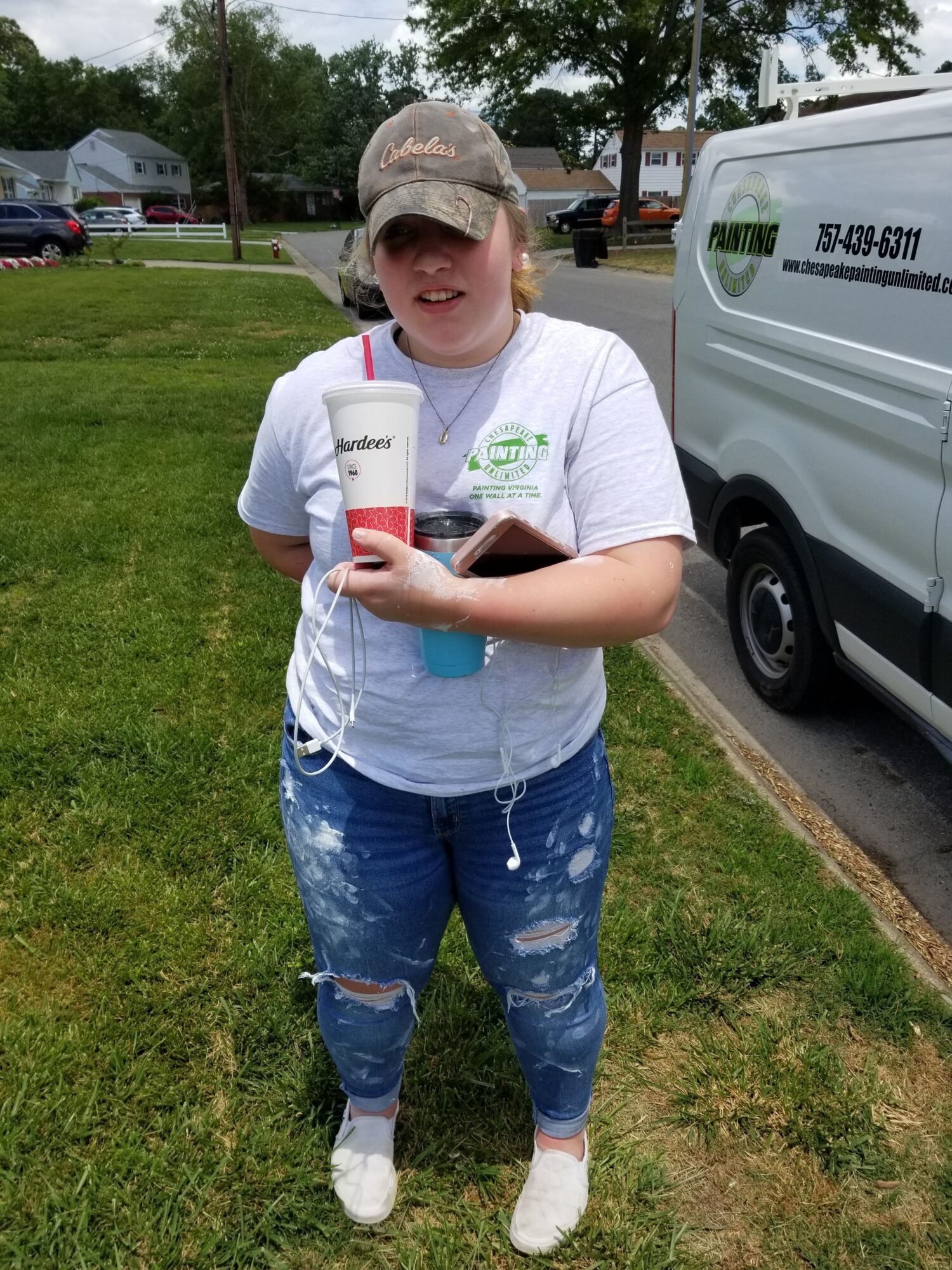 Young person with cap holding a drink cup, standing on grass near parked vehicles, wearing white shirt and jeans.