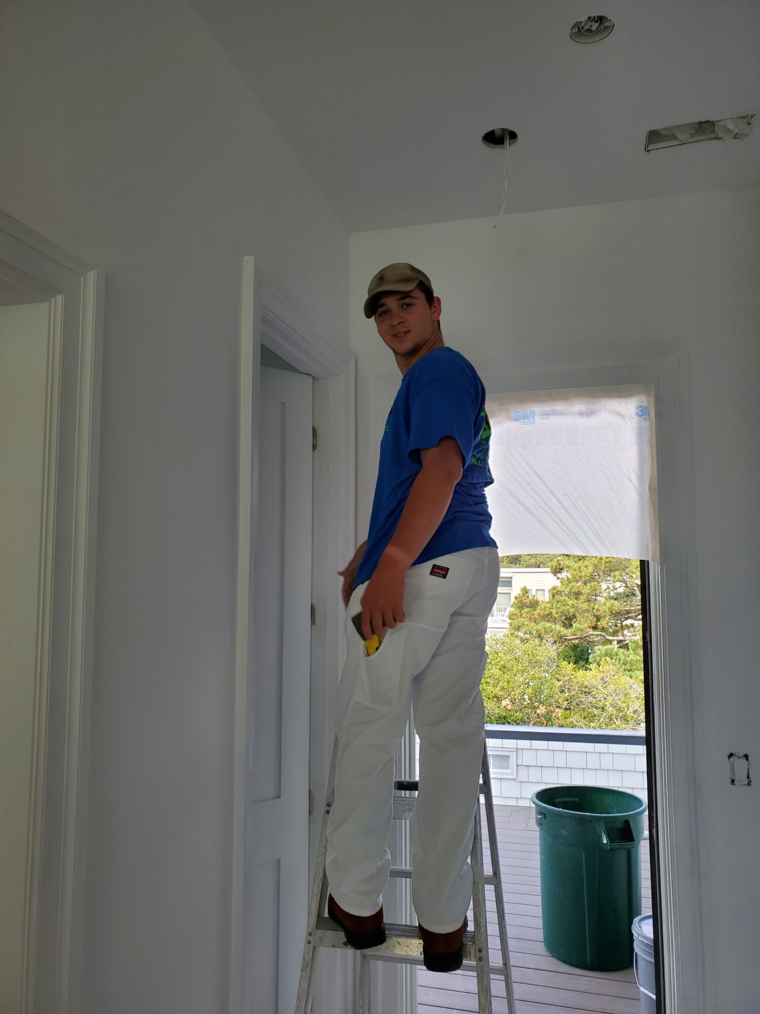 Person standing in doorway, wearing blue shirt and white pants, with a window and green trash bin outside.