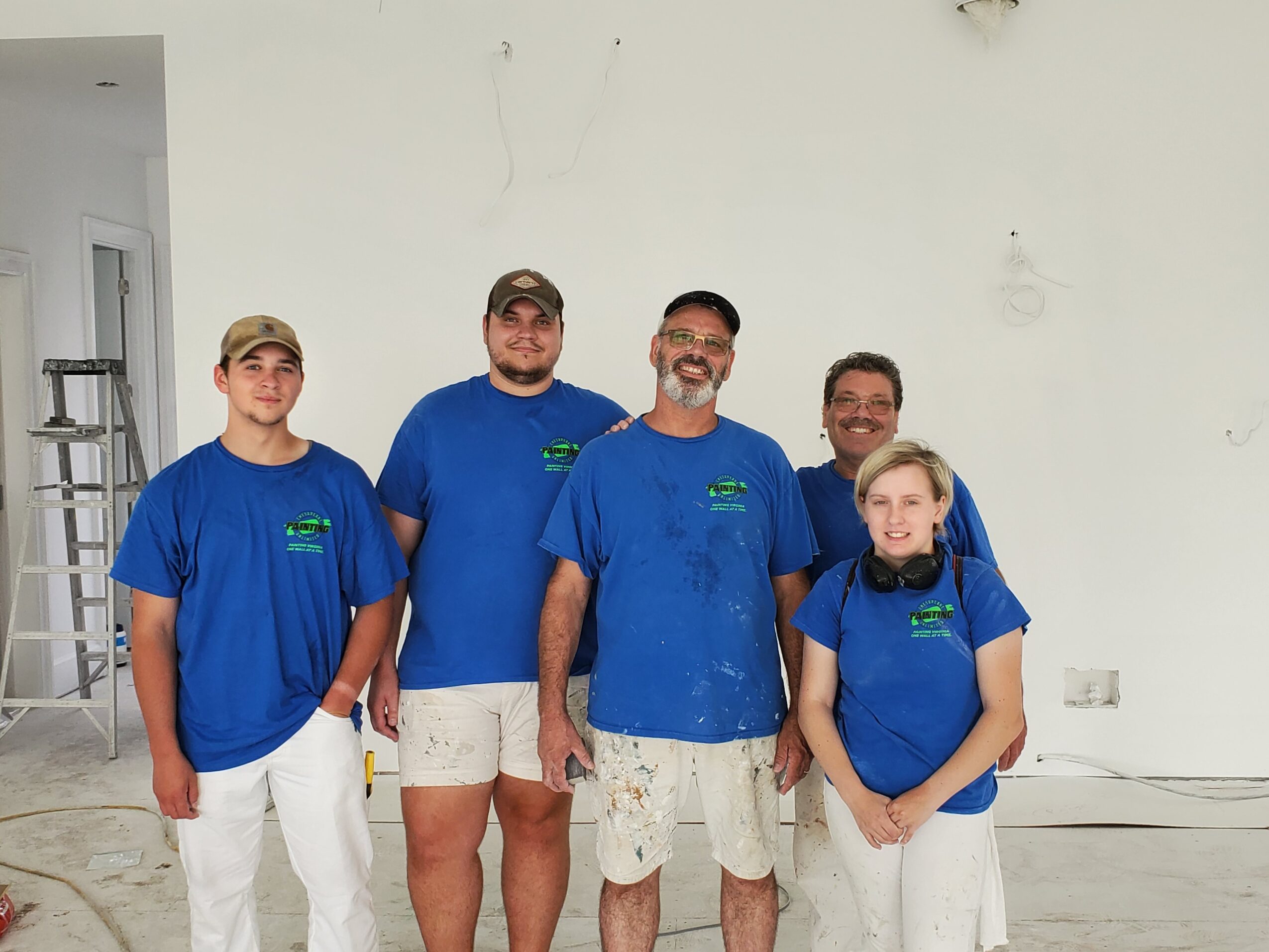 Five people wearing blue shirts and white shorts or pants standing indoors against a plain wall.