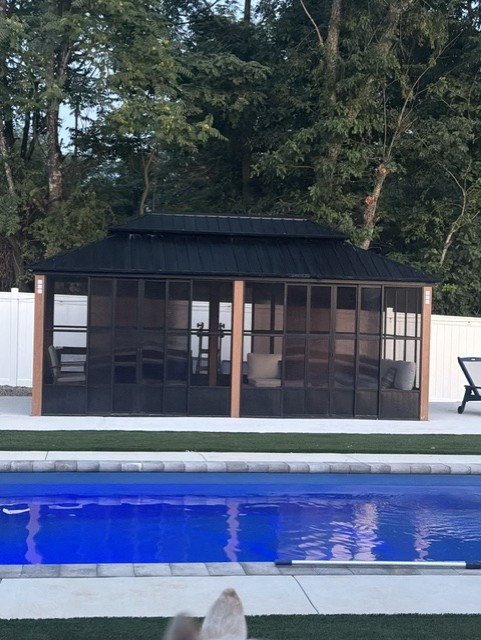 A screened outdoor structure with a black roof, situated behind a white fence, next to a swimming pool with blue water.