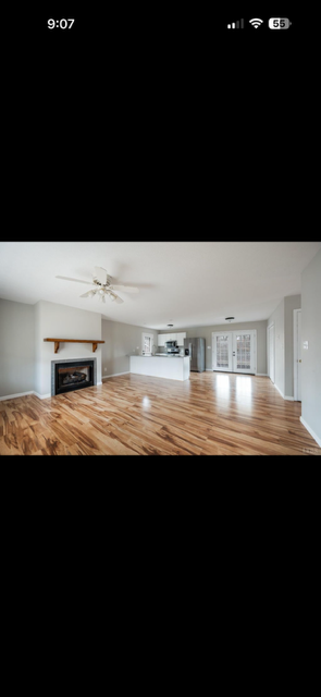 Empty living room with hardwood floors, fireplace, ceiling fan, and large windows, bright and spacious.