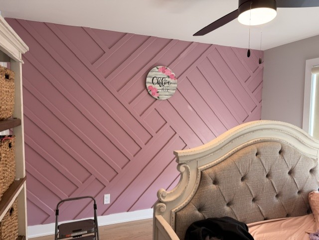 Bedroom corner with pink diagonal wall, tufted headboard, and a round wall clock, ceiling fan, and window visible.