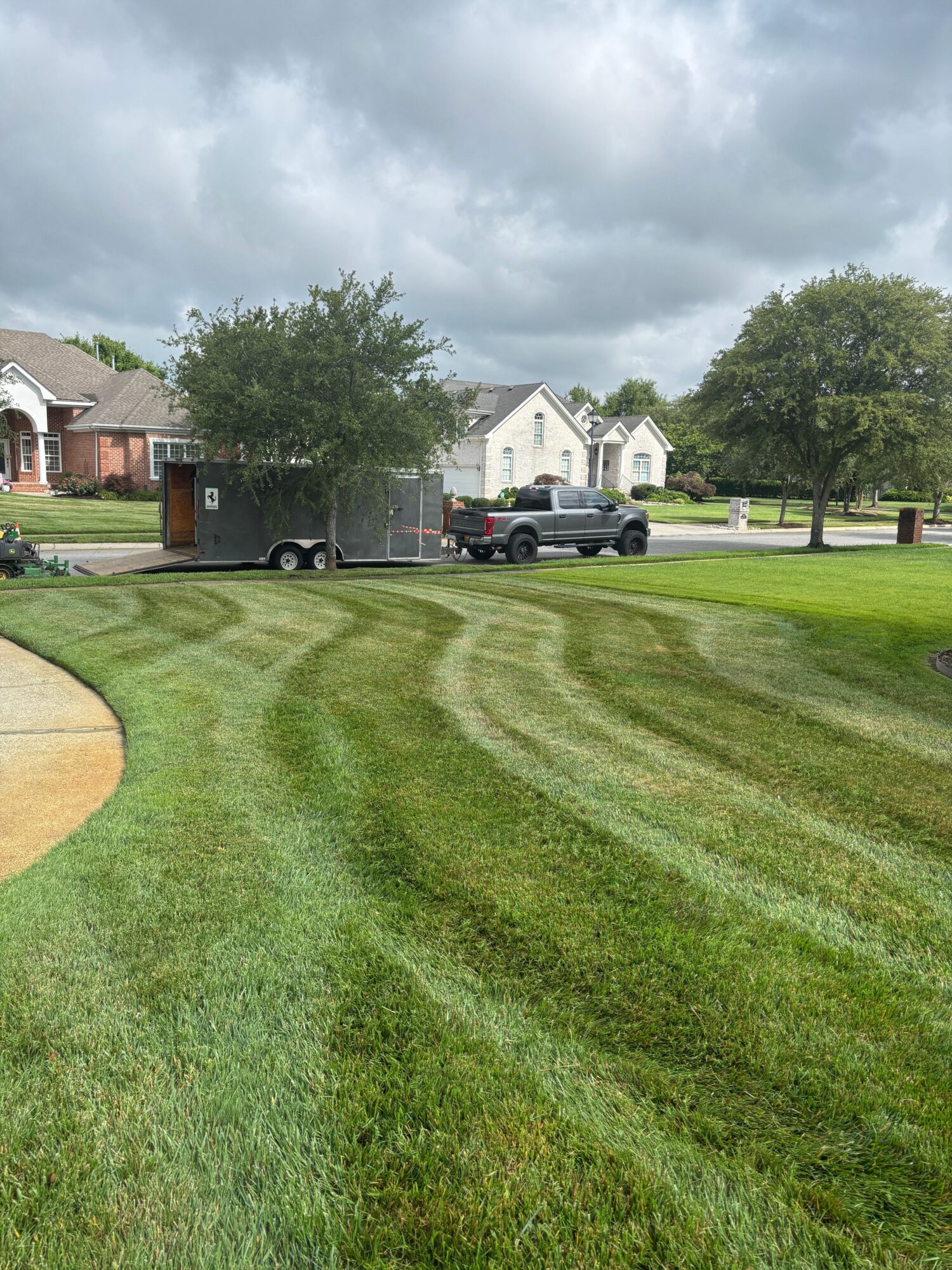 A grassy area with a small sand patch, trees, houses, and parked cars under cloudy sky.