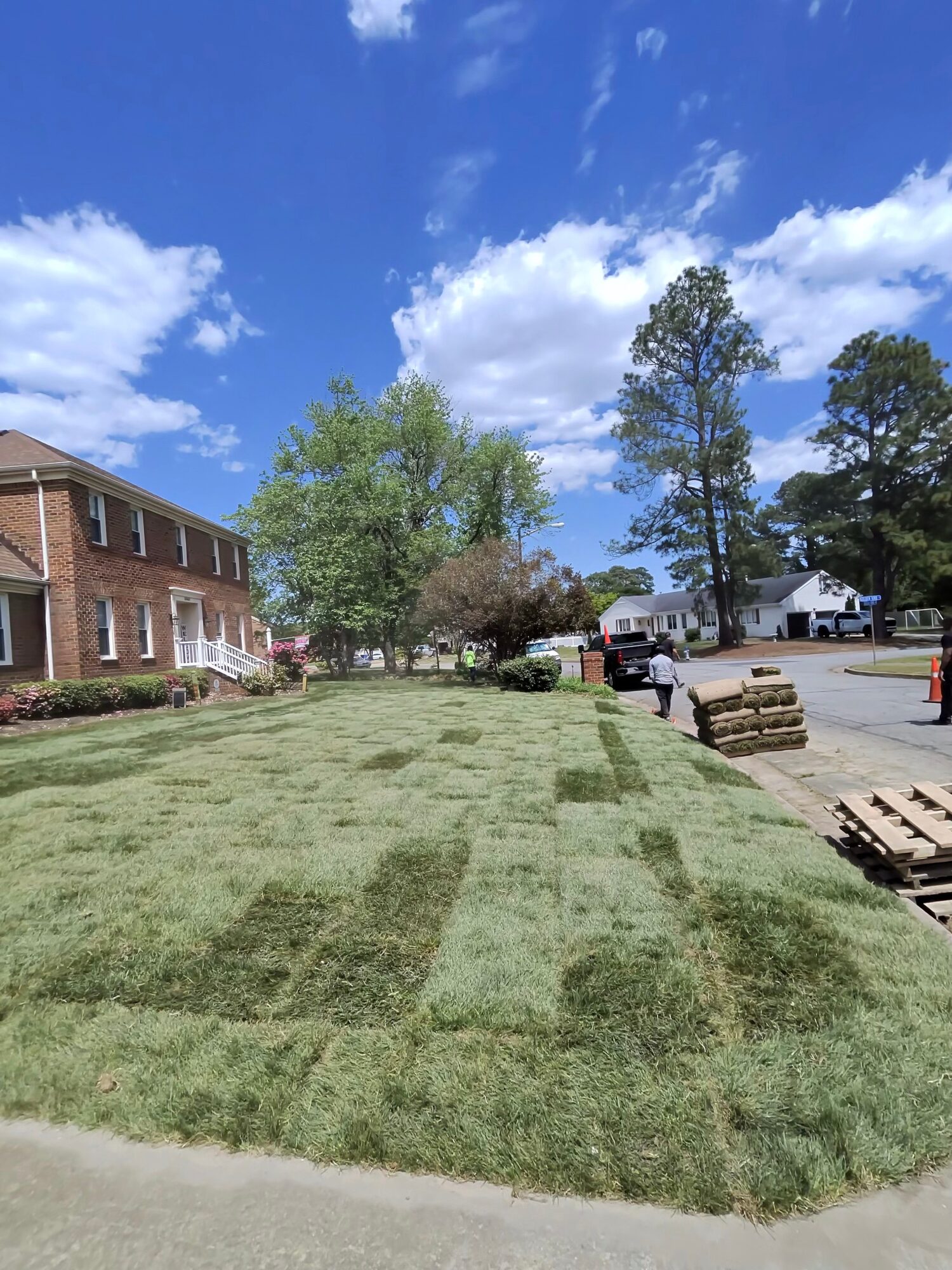 A grassy area with trees, a brick building on the left, and a parking lot with vehicles and pallets on the right under a blue sky with clouds.