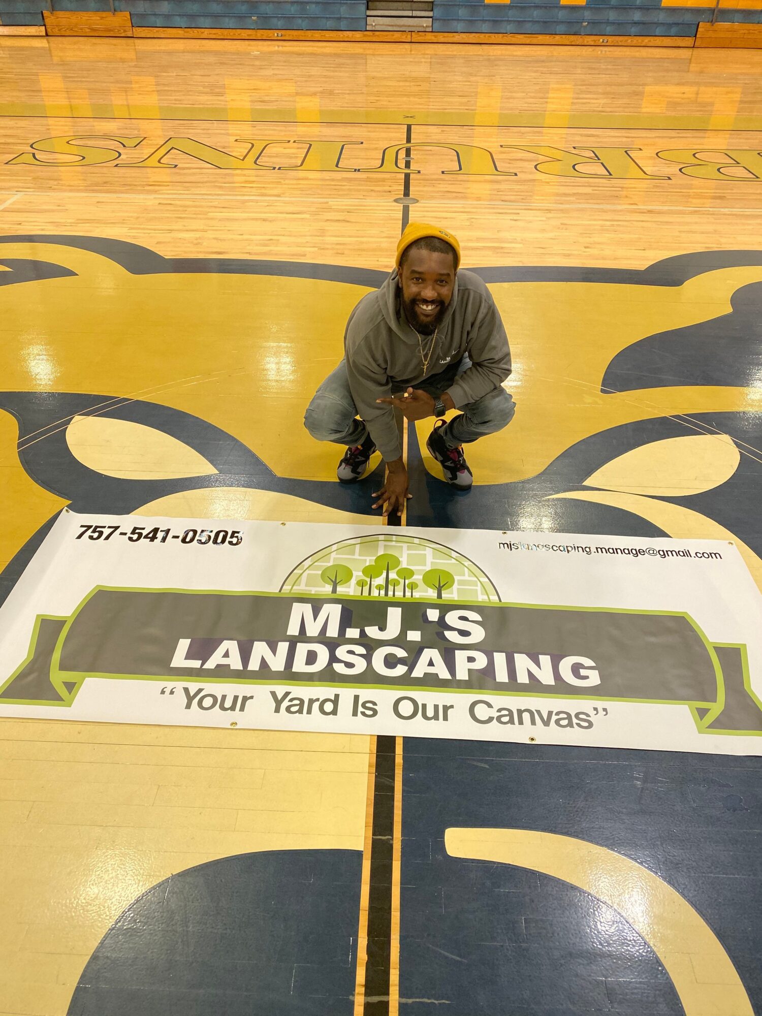 Person crouching next to a landscape sign on a gymnasium floor, with a yellow and black design, holding a camera.