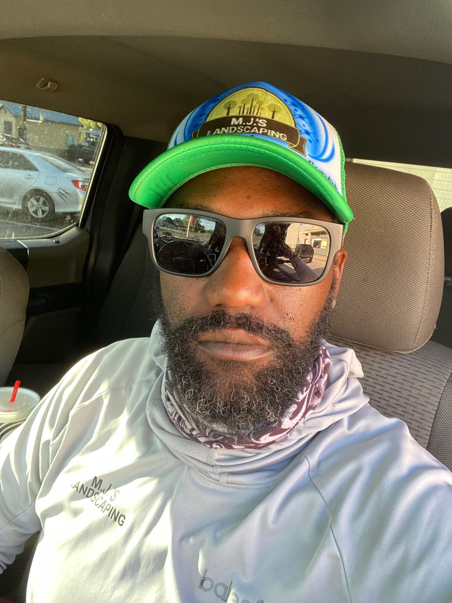 Man with sunglasses and beard wearing a cap and white shirt, sitting in a vehicle, with a window showing a street scene.
