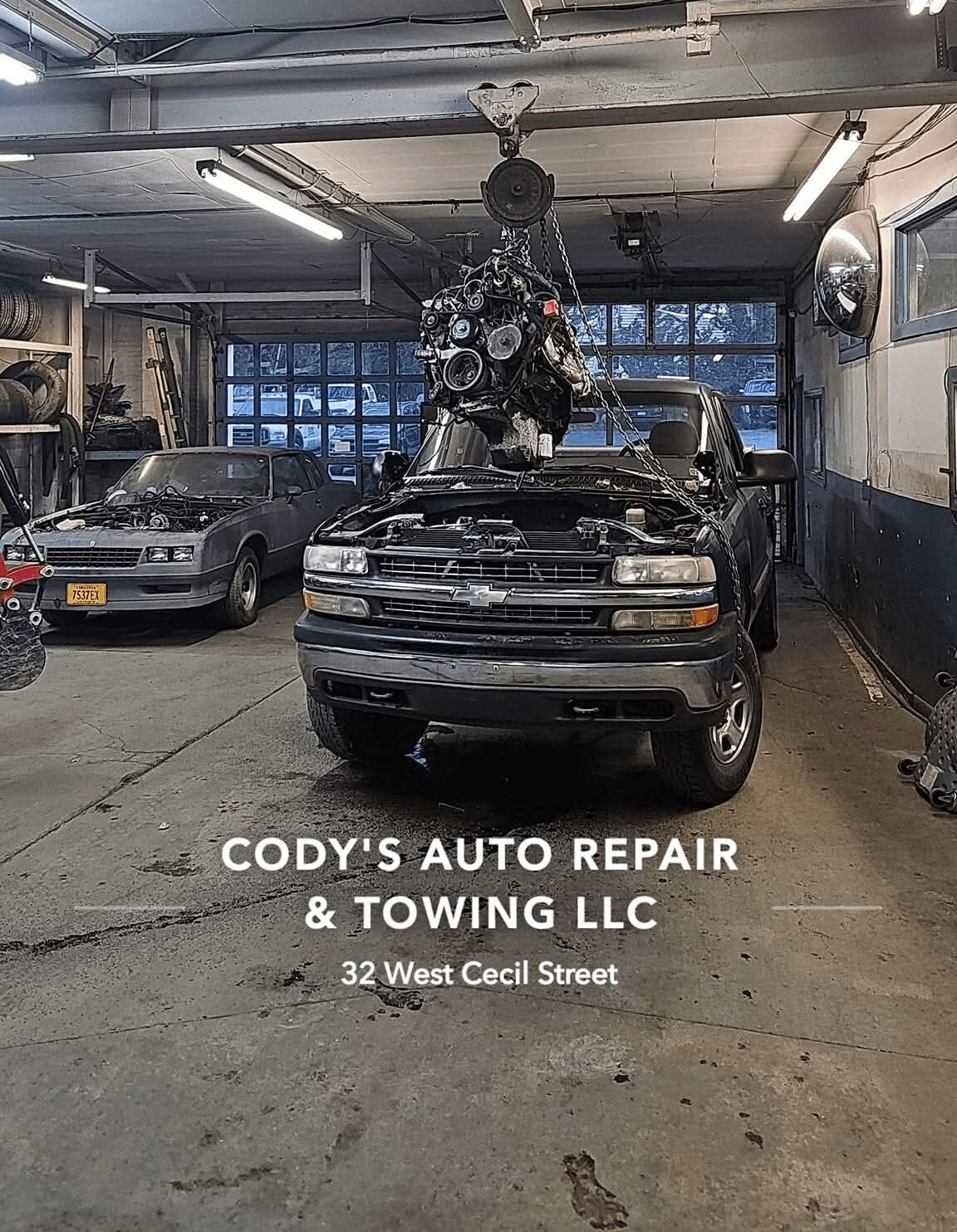 Car inside repair shop with engine hoisted above hood, two other cars visible, workshop with tools and windows in background.